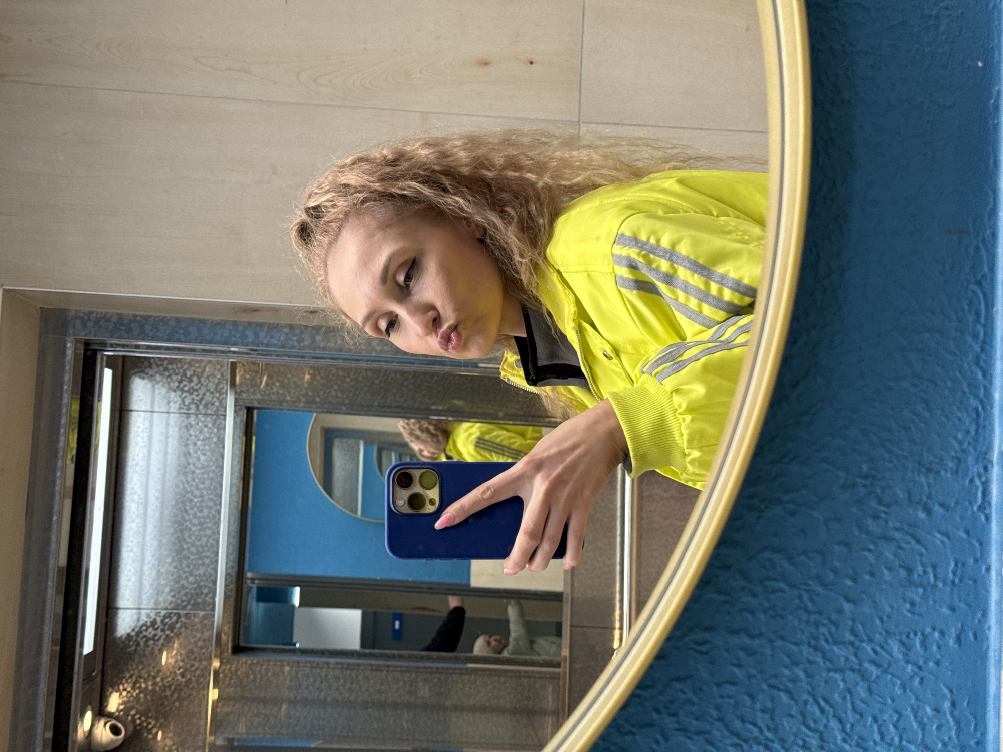 A young woman with light blonde, curly hair is taking a selfie in a bathroom mirror, likely in the city of Perm, Russia. She is wearing a bright yellow jacket with grey stripes on the sleeves. Her phone, a blue iPhone with multiple camera lenses, is visible. The reflection in the mirror shows the bathroom interior, including what appears to be a changing table, and partially visible individuals. The mirror itself has a blue, textured frame with a gold trim. The background includes a light-colored wall and the metal framework surrounding the mirror. The woman is making a "duck face" expression.