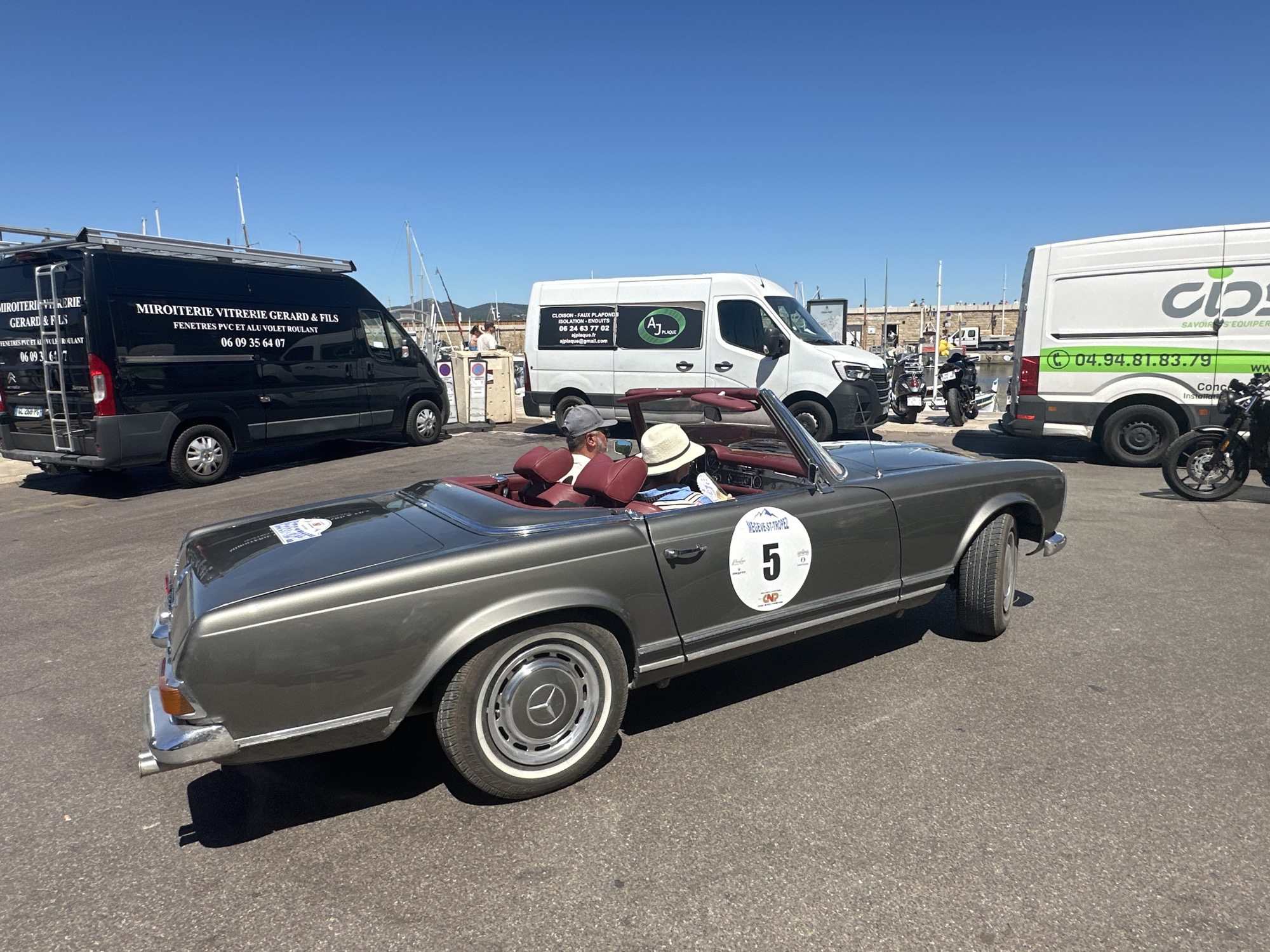 A grey Mercedes-Benz SL convertible with a red interior is parked on an asphalt surface. Two people, a male wearing a grey baseball cap and a female wearing a straw hat, are seated in the convertible. A white circular sticker with the number "5" in black is affixed to the car's door, along with other text and logos. Behind the convertible, two white vans and a black van are parked. The white vans have various company names and phone numbers printed on them. The black van has "MIROITERIE VITRERIE GERARD & FILS" printed on its side. Several motorcycles are visible parked behind the white vans. The scene appears to be in a port or marina area, with boats and masts visible in the background under a clear blue sky. This location is identified by the text on the sticker as MEGÈVE-ST-TROPEZ, indicating the event likely involves a journey or connection between these two locations.