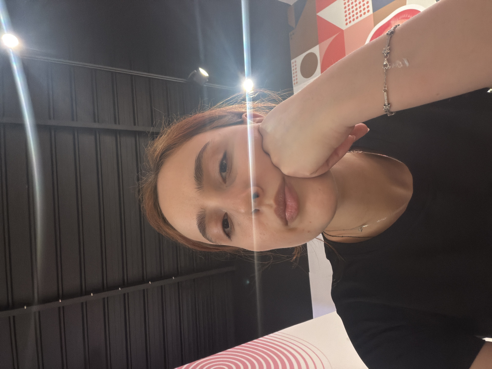 This is a close-up, head-and-shoulders shot of a young woman with auburn hair, resting her chin on her hand. She is wearing a black t-shirt and a silver bracelet adorned with small charms, one of which appears to be a lowercase "love" charm. A silver necklace is also visible. The background consists of a dark, slatted ceiling with lighting fixtures and a colorful geometric pattern on a wall to the right. To the left, a portion of a pink and white spiral design is visible. The lighting in the image is characterized by a strong glare from overhead lights. No information about the location in Strasbourg, France, can be determined from the visual content of the image.