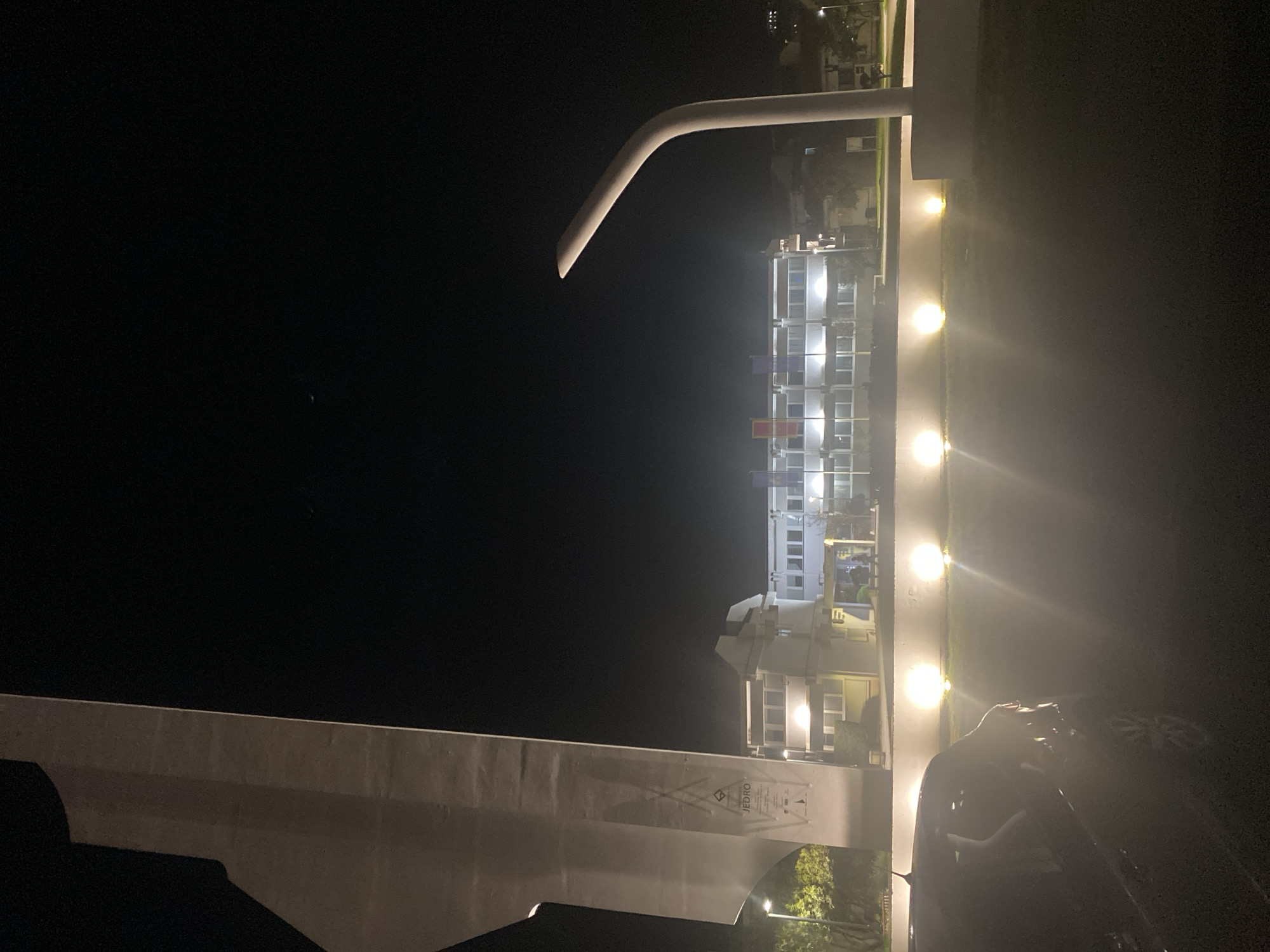 A night scene in Budva, Montenegro shows a building with illuminated windows, including some with colored curtains. A white railing and a curved metal pipe are visible. A car is parked in the foreground, with the outline of a person beside it. Trees are seen near the building. String lights line the edge of a structure next to the building. A sign is affixed to a wall. The sky is dark.