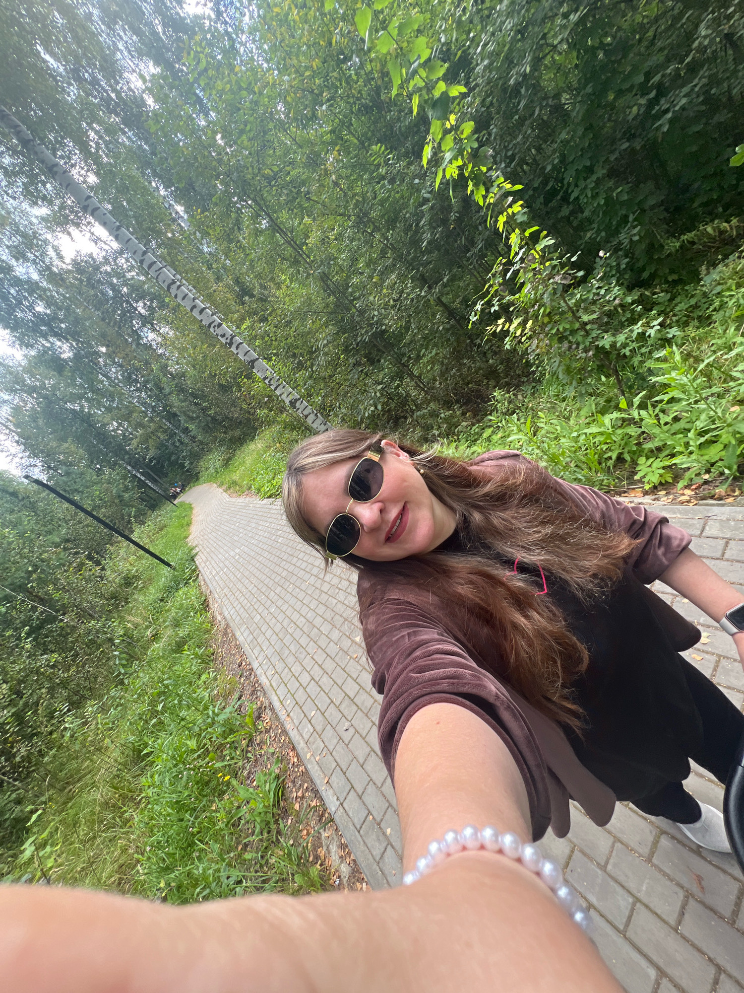 A young woman with long, brown hair and sunglasses takes a selfie outdoors. She is wearing a brown velvet hoodie over a dark shirt and dark pants. A pearl bracelet adorns her left wrist. She is standing on a paved walkway lined with trees and grass. In the distance, two people can be seen walking or cycling on the path. The setting appears to be a park or wooded area. The image does not provide specific geographical markers to pinpoint the location in Noviy, Russia.