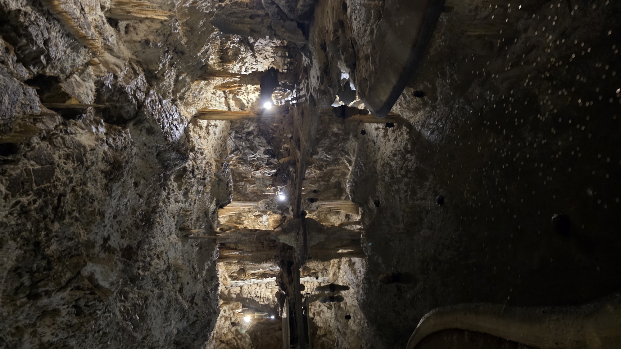 The image depicts the interior of a cave in Vavřinec, Czechia. The scene is a view looking upwards into a cavern with rough, textured rock formations on either side. Stalactites hang from the ceiling and in some areas, wooden beams appear to be embedded in the rock, possibly for structural support or as part of an exhibition.
Artificial lights, some appearing as spotlights, are strategically placed within the cave, illuminating the formations and creating areas of light and shadow. These lights highlight the rugged texture and layered nature of the rock. On the right side of the cavern, the ceiling is darker and dotted with numerous small, bright circular reflections, possibly from coins that have been thrown or affixed to the surface. There are also a few darker, spherical objects attached to this section of the wall.
The overall impression is of a natural cave that has been adapted for public access, likely as a tourist attraction. The lighting suggests it is either perpetually dimly lit or illuminated for viewing at any time. The presence of coins suggests a tradition or belief associated with the location. The image conveys a sense of wonder and exploration of an underground natural environment. No people are visible, nor is any text apparent. The weather and time of day are irrelevant given the enclosed underground setting.