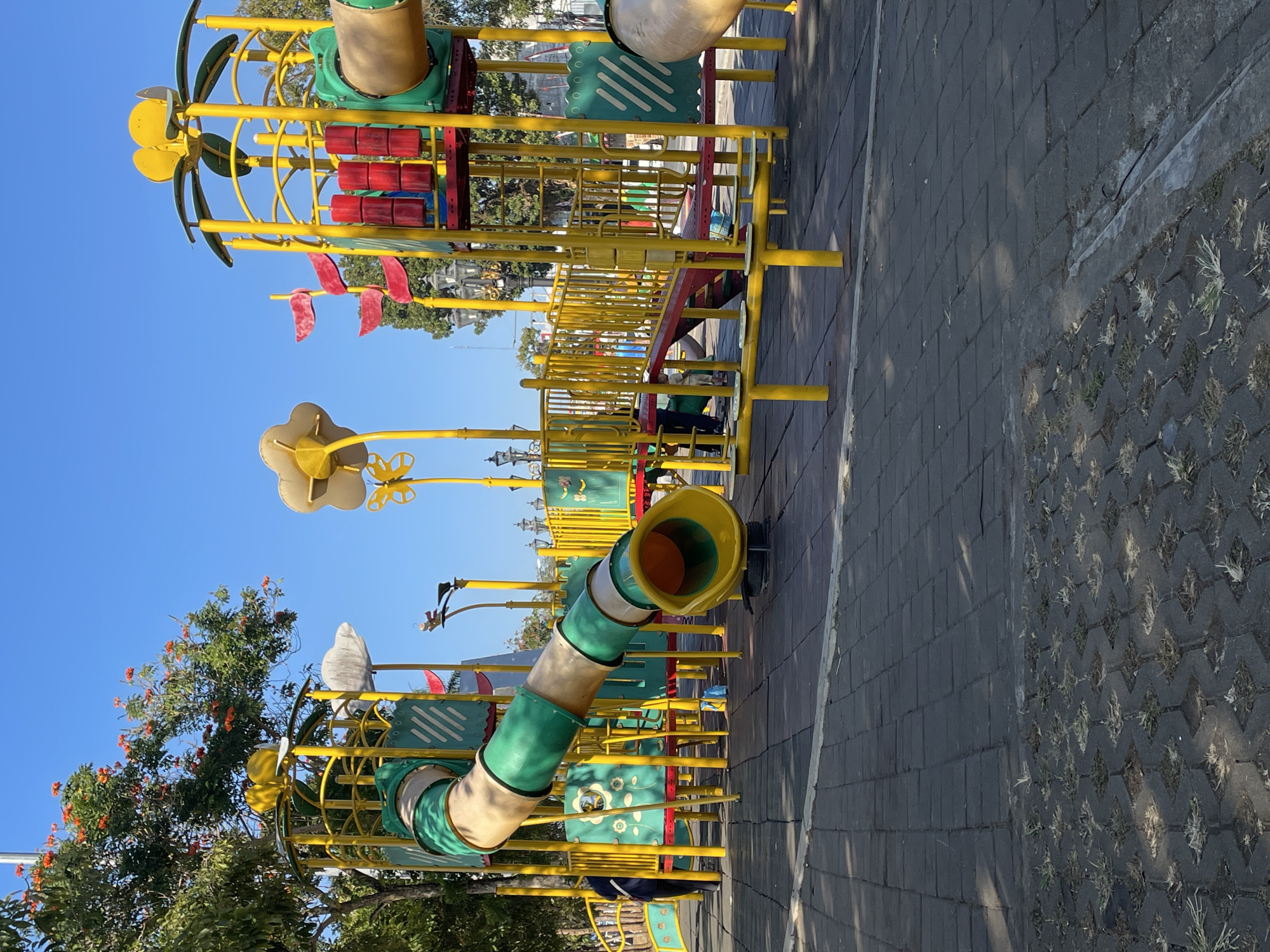 This is a full-shot, eye-level photograph of a brightly colored children's playground located in the city of Purworejo, Indonesia. The playground features a large, multi-level structure made of yellow metal poles with green and beige tubular slides and a red ramp. Decorative yellow flower-shaped elements are attached to the top of some of the poles. The ground is paved with dark gray interlocking tiles, with a patch of patterned concrete with grass in between. Several children, some wearing green and others darker clothing, are visible playing on the structure. A person in dark clothing with a white strap is standing near the left side of the playground. The sky above is clear and blue. Trees with orange flowers and green leaves are visible on the left and in the background, along with some decorative lampposts. The lighting suggests it is daytime.