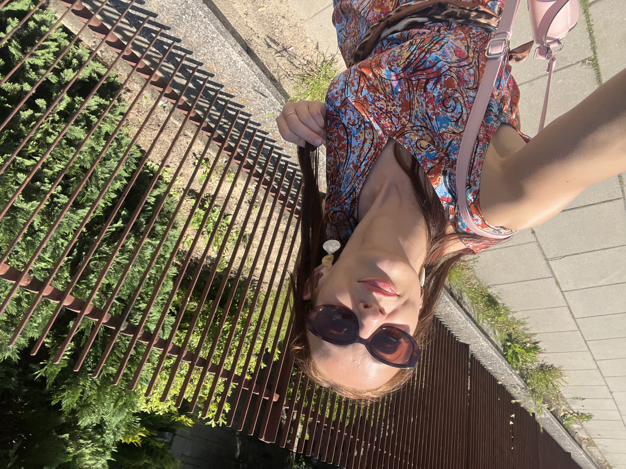 A woman is captured in a selfie, taken outdoors during the day. She is wearing a colorful, patterned dress with shades of red, blue, and orange. A light pink crossbody bag with a strap is visible over her shoulder. She has on large, tortoiseshell sunglasses and prominent, dangling earrings. Her hand is extended, holding a lock of her long, dark brown hair. A thin gold ring is on her finger. The background features a rusty metal fence, green bushes, and a paved walkway or sidewalk. The lighting suggests a sunny day, casting shadows from the fence. The woman's pose and direct gaze into the camera indicate she is actively taking the photograph. The overall impression is casual and summery.