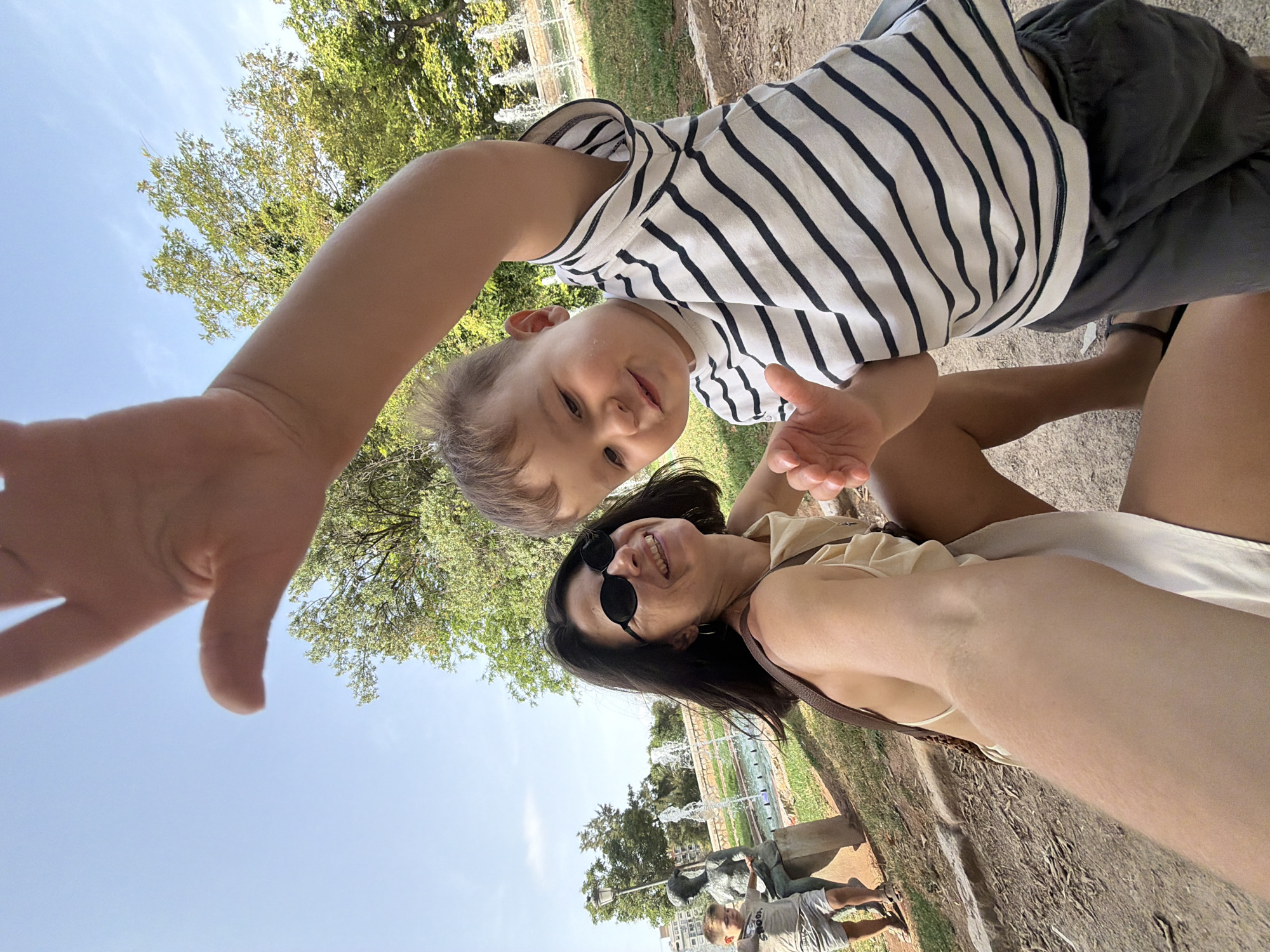 This media file is a selfie taken by a woman with a young child in an outdoor park setting. The woman is in the foreground, wearing sunglasses and a light-colored top. The child is positioned above her, looking towards the camera with a smiling expression. The child is wearing a striped shirt and dark shorts. In the background, there are trees, a fountain with water jets, and some structures that suggest an urban park. The sky is blue, indicating it is daytime and likely clear weather. The overall mood appears joyful and casual, as suggested by the smiles of both subjects and the informal nature of a selfie. The photo is oriented sideways. There is no visible text in the image.