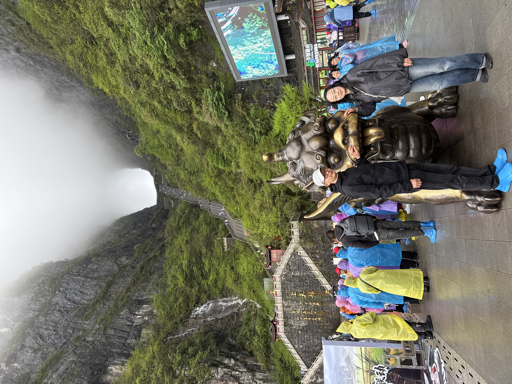 The photograph depicts the Tianmen Mountain Scenic Area in Zhangjiajie, China, featuring various elements and individuals. In the foreground, two people are posing next to a bronze statue of a mythical creature. Both are wearing shoe covers. A line of individuals clad in colorful rain ponchos stands nearby. To the upper right, one can see a digital screen displaying vibrant imagery. The background features a mountainous landscape covered in lush greenery. A staircase is visible, winding up the mountainside, and a waterfall cascades down the rocks. The summit of the mountain is shrouded in mist, creating a dramatic effect.