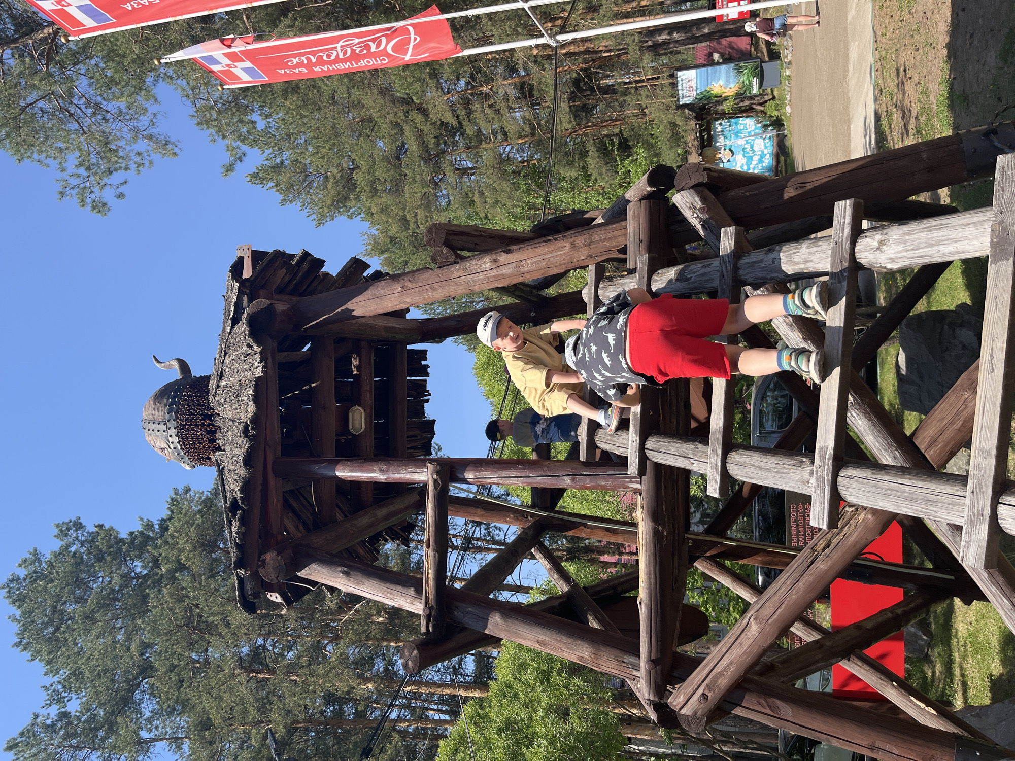 A wooden structure reminiscent of a historical tower is situated in a park-like setting in Chaykovsky, Russia. Two children are climbing on the structure, one wearing a yellow shirt and a light-colored hat, and the other dressed in red shorts and a patterned shirt. Above the structure, a red banner with white Cyrillic text flutters against a clear blue sky, with pine trees in the background. The tower itself is constructed from dark wooden beams and features a decorative, possibly metal, dome on top. Additional signage and people are visible in the background, suggesting a public space.
