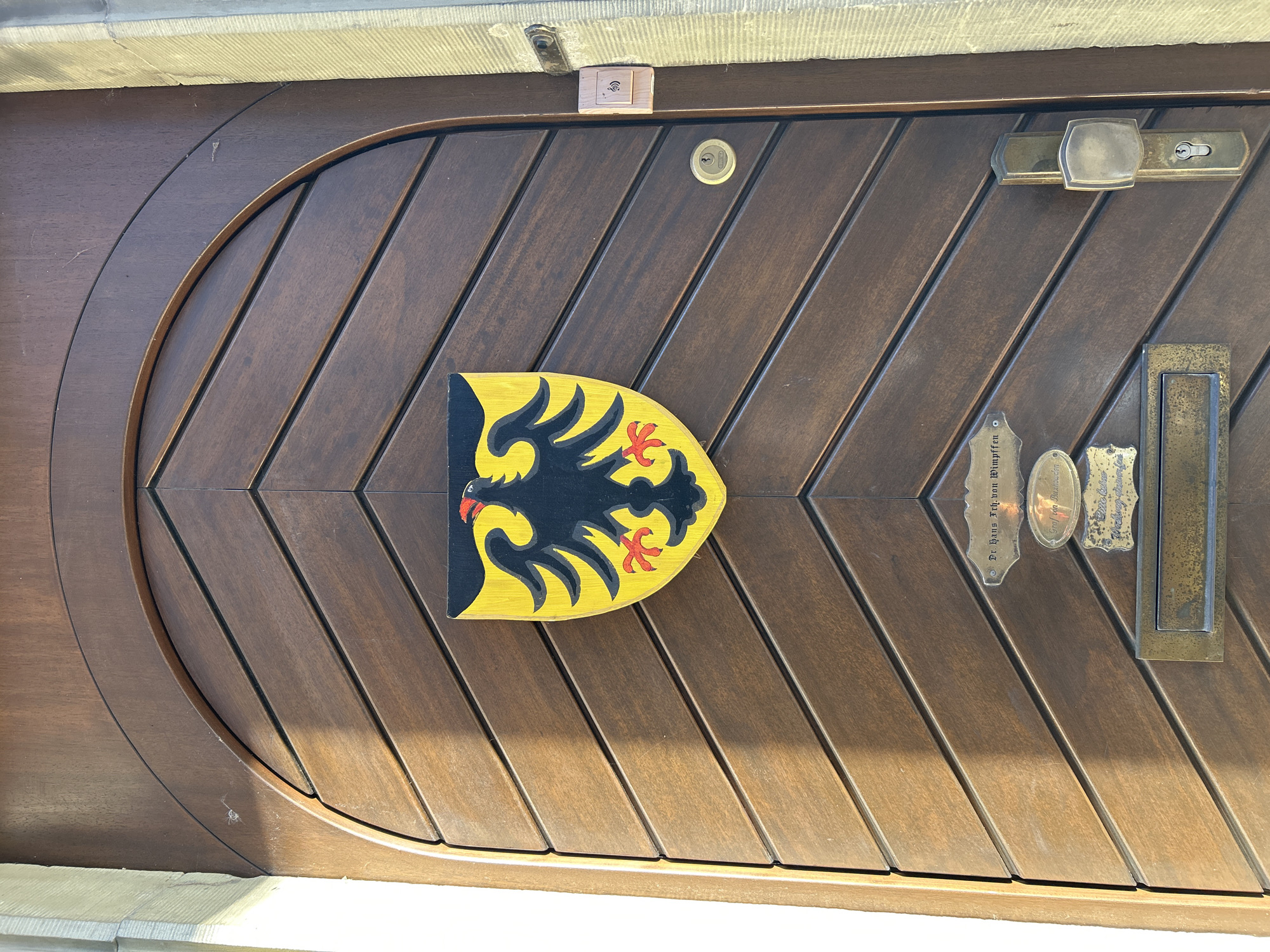 A photograph shows the front door of a building in Unknown, Unknown. The door is made of dark brown wood and features a chevron pattern. A yellow and black shield with a red claw emblem is centrally mounted on the door. Various metal fixtures including a door handle, keyhole, letterbox, and inscribed nameplates are visible on the right side of the door. A small electronic device is affixed above the door, near the top of the frame. The building's exterior is a light-colored stone.