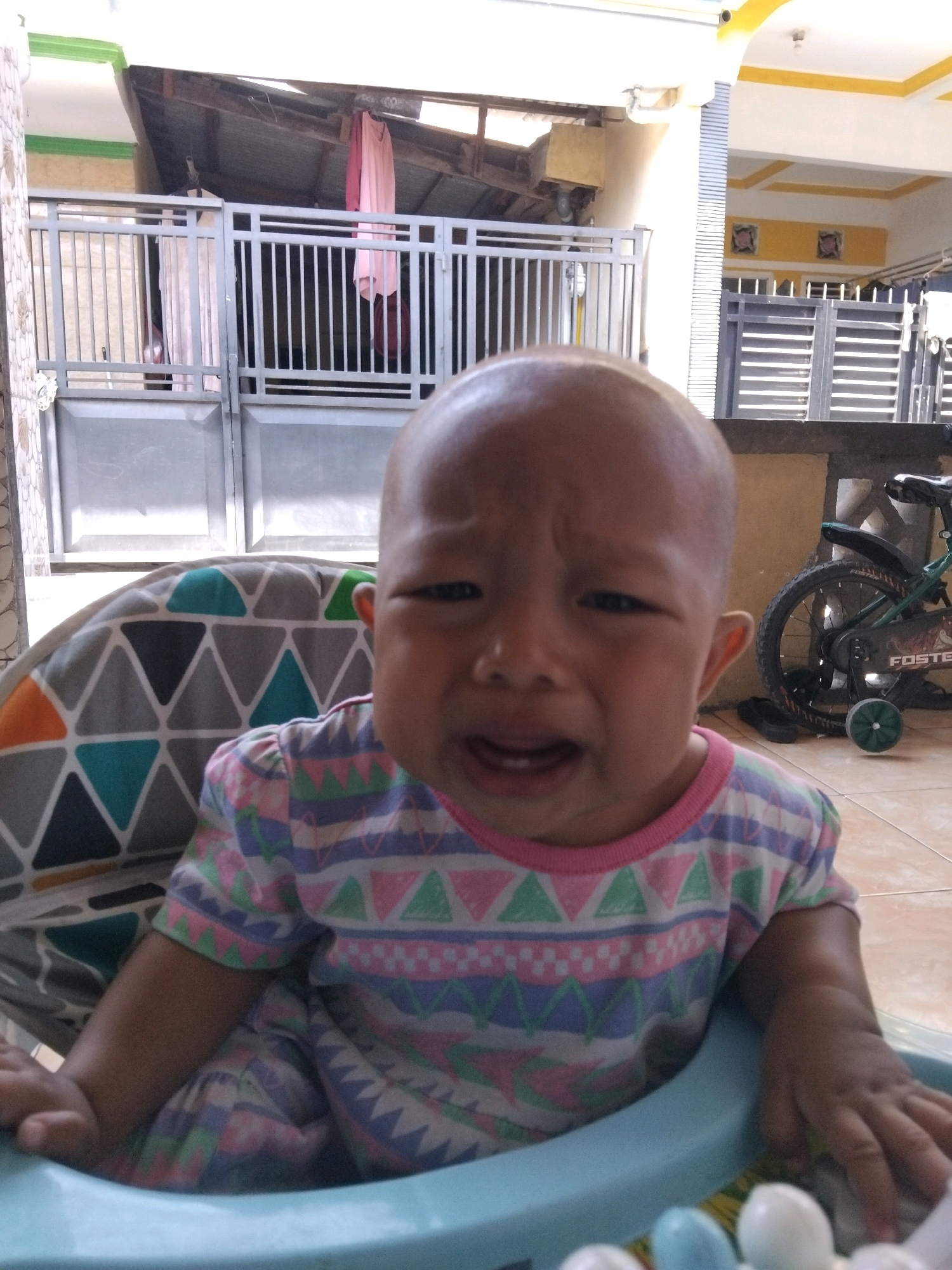 A bald baby wearing a colorful patterned shirt is sitting in a high chair. The baby has a distressed expression and is crying. The high chair is decorated with geometric patterns in gray, teal, and orange. In the background, there is a metal gate, a bicycle, and part of a building with a yellow and white facade. The scene appears to be outdoors, possibly on a patio or in a yard. The location is identified as Kepuh Kiriman, Indonesia.