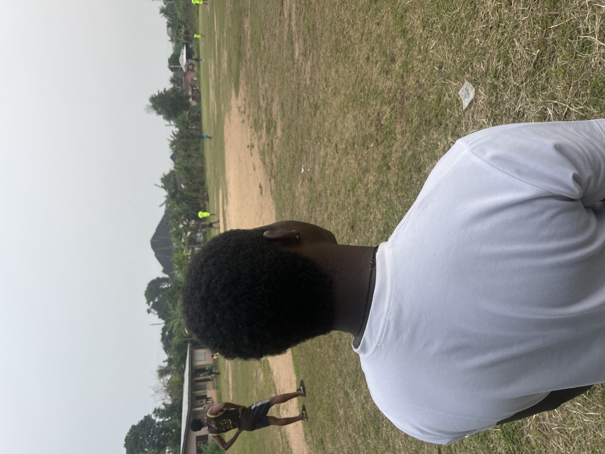 A person is wearing a white shirt in an outdoor setting. The background features a field with patches of grass and bare earth. In the distance, there are trees and what appears to be a building or structures. Another person wearing a dark athletic uniform with a yellow number is walking or stretching in the foreground. A piece of white trash is on the ground. The exact location in Ikot Ekpene, Nigeria, is not discernible from the image.