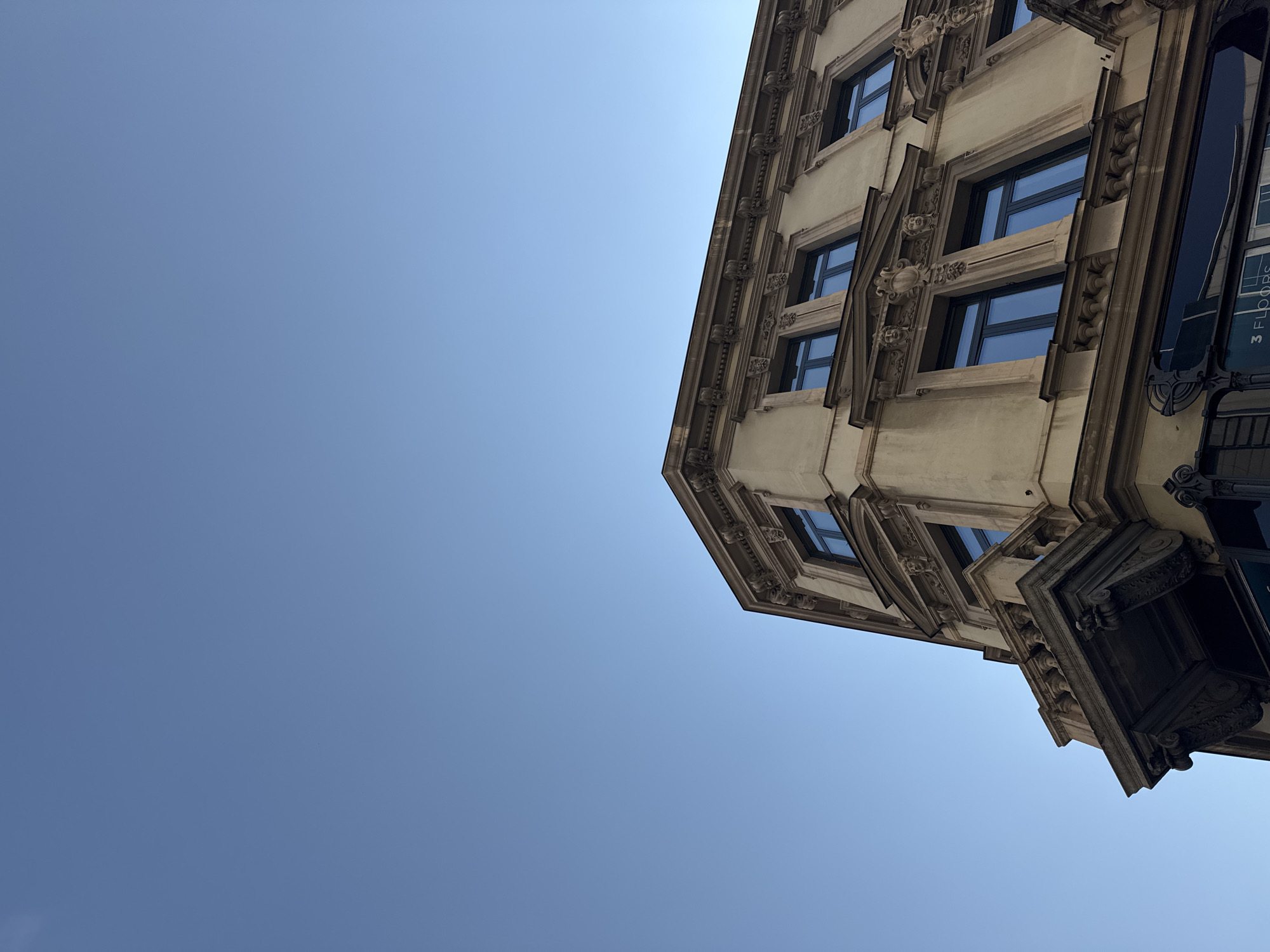 This is a low-angle, eye-level shot of a tan building in Belgrade, Serbia. The building has multiple stories with ornate architectural details, including detailed moldings and carvings above windows. The windows themselves are dark blue. The sky is a clear, bright blue. The image appears to have been taken during daylight. There are no people visible in the frame.