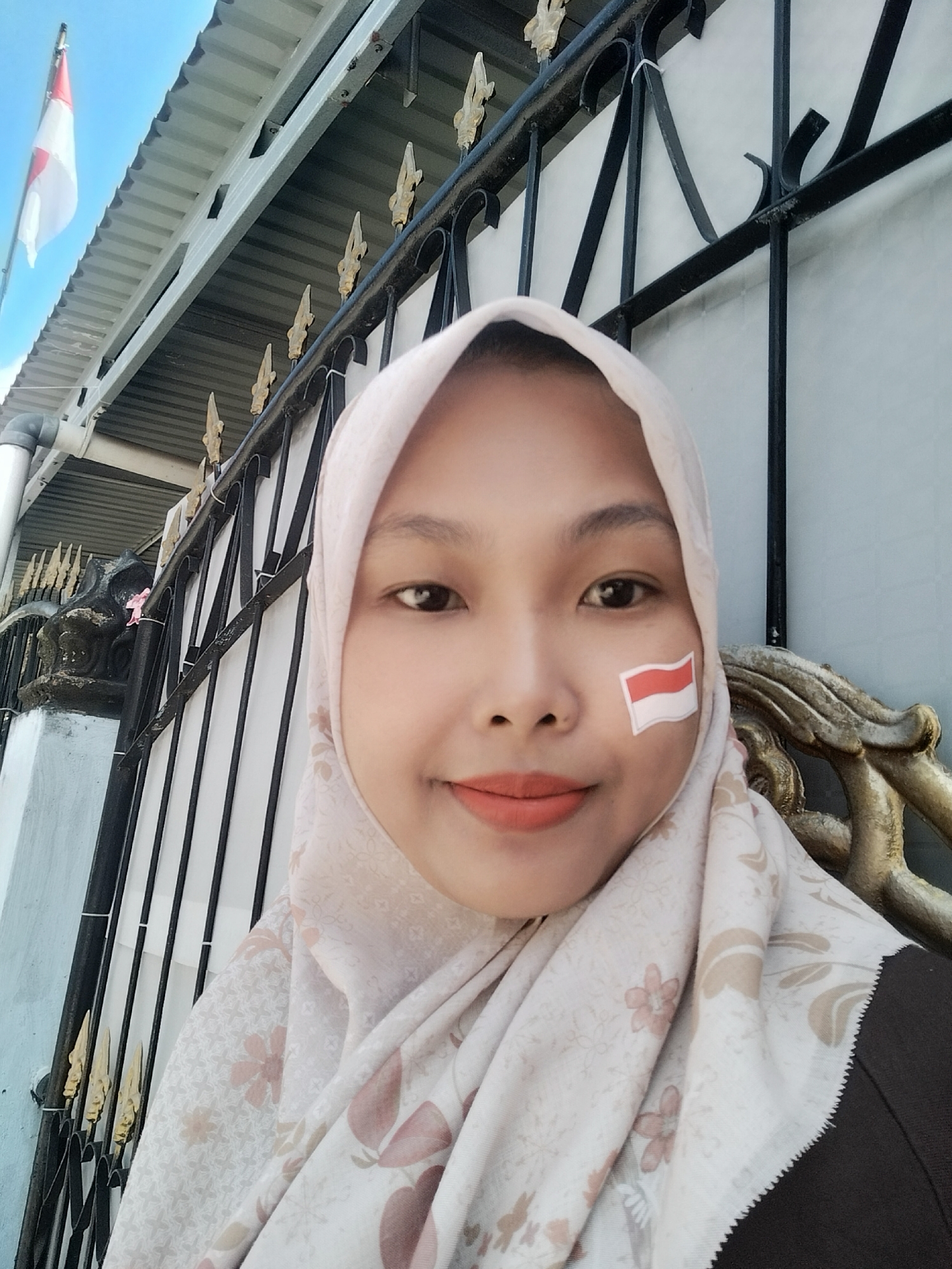 A young woman wearing a hijab and a light-colored scarf with a floral pattern is pictured in a close-up, forward-facing selfie. A small Indonesian flag is adhered to her right cheek. Her lips are painted with a peachy-orange lipstick. The background includes a black metal fence with decorative golden elements, a light blue wall, and a glimpse of a white building with a corrugated roof and a flagpole with the Indonesian flag. The scene appears to be outdoors.