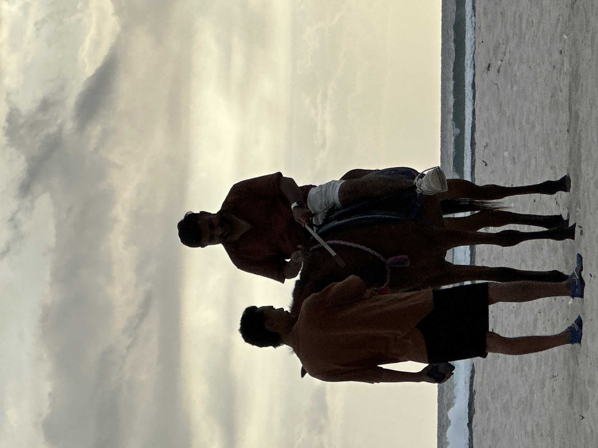 A medium shot depicts a man riding a horse on a beach next to the ocean in the city of Unknown, Indonesia. Another man is standing next to the horse. The lighting suggests it is dusk or dawn. The sky is cloudy. The rider is wearing a maroon shirt and white shorts, and the man next to the horse is wearing a brown shirt and black shorts. Both men appear to have dark hair.