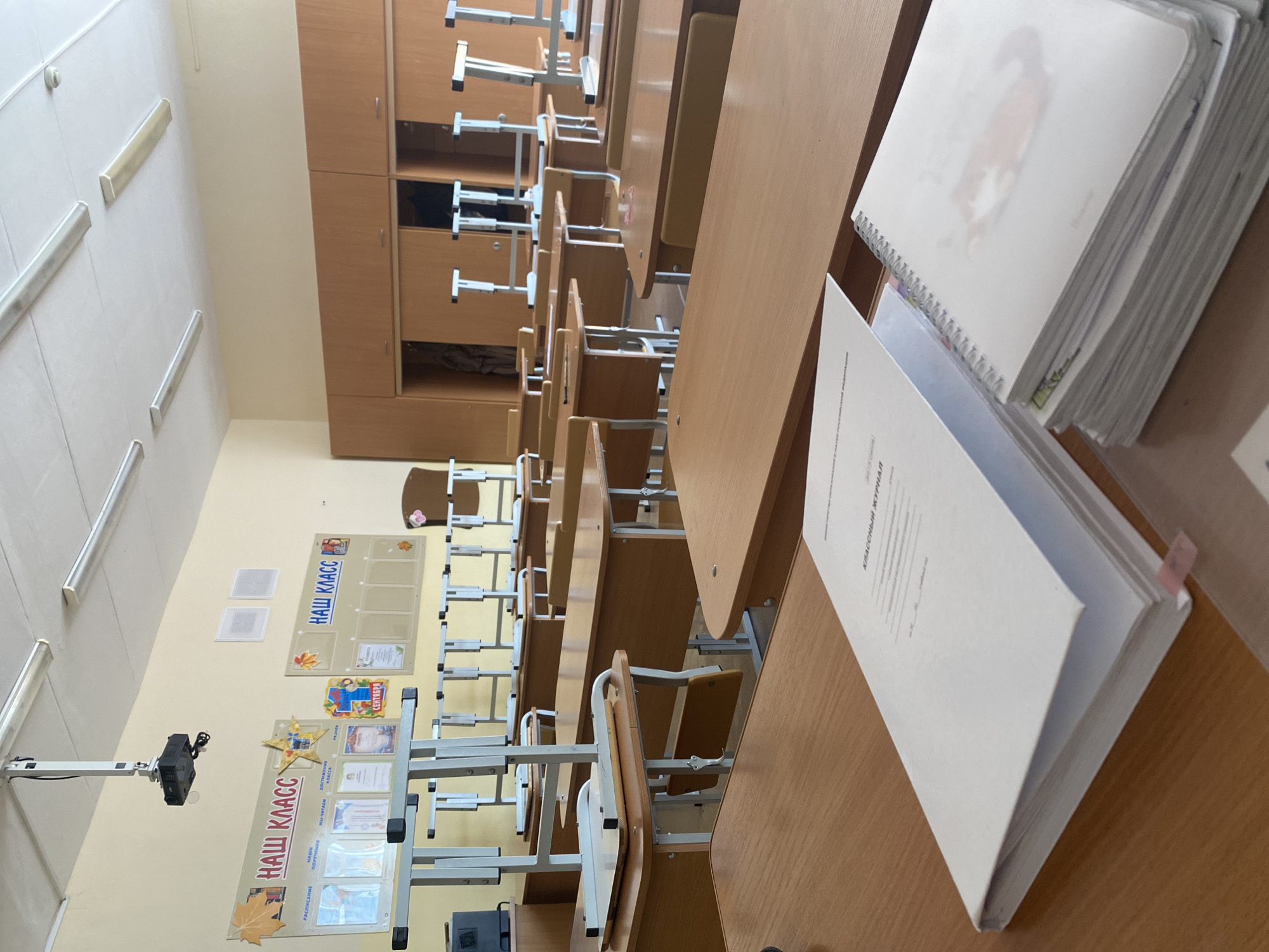 The image shows an empty classroom, likely in a school, in Chaikovsky, Russia. Rows of wooden desks and chairs occupy the foreground and middle ground. A large, light-colored cabinet spans the wall on the left. On the wall are posters labeled "НАШ КЛАСС" (Our Class), suggesting it's a primary school classroom. A projector is mounted on the wall. A stack of notebooks and a document are on a desk in the foreground. The environment is sterile and devoid of people.