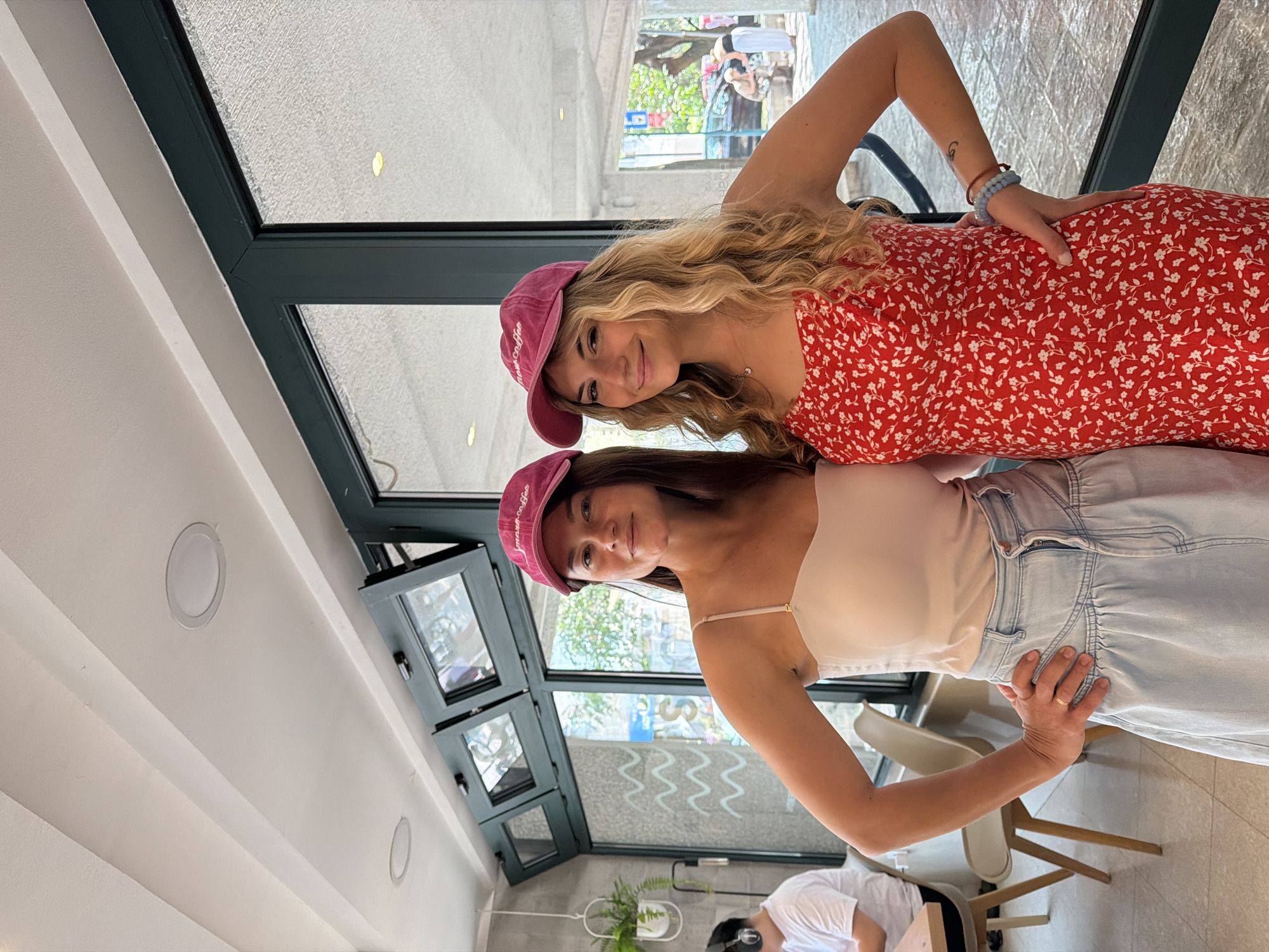Two smiling women wearing matching pink caps are posing in front of a window. The woman on top has long blonde wavy hair and is wearing a red floral dress. The woman below has brown hair and is wearing a white top and a light blue denim skirt. Both women have their hands on their hips.
Through the window, a street scene is visible. The scene appears to be indoors in a well-lit space. There is a ceiling with recessed lighting and the frame of the window is a dark greenish-gray color.
Based on visual cues such as architectural style and vegetation, the location appears to be in a coastal European city. Given the presence of a sign, the location may be in Budva, Montenegro.
