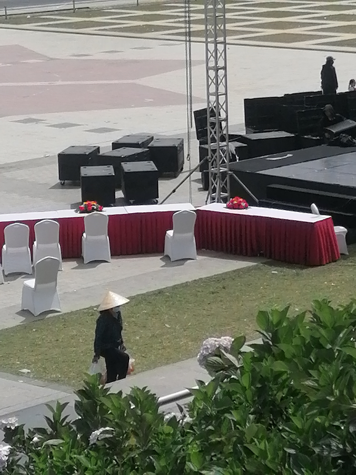 A lone individual wearing a conical hat and a face mask walks on a grassy area in Da Lat, Vietnam. The person is dressed in dark clothing and is carrying plastic bags. In the background, there are several white chairs arranged around a long table covered with a red cloth. Further back, a large stage structure with black boxes is visible, along with a metal tower. The ground in front of the stage is paved with interlocking squares. In the distance, there are more paved areas and faint criss-cross markings on a lighter surface. The scene appears to be set up for an outdoor event or performance.