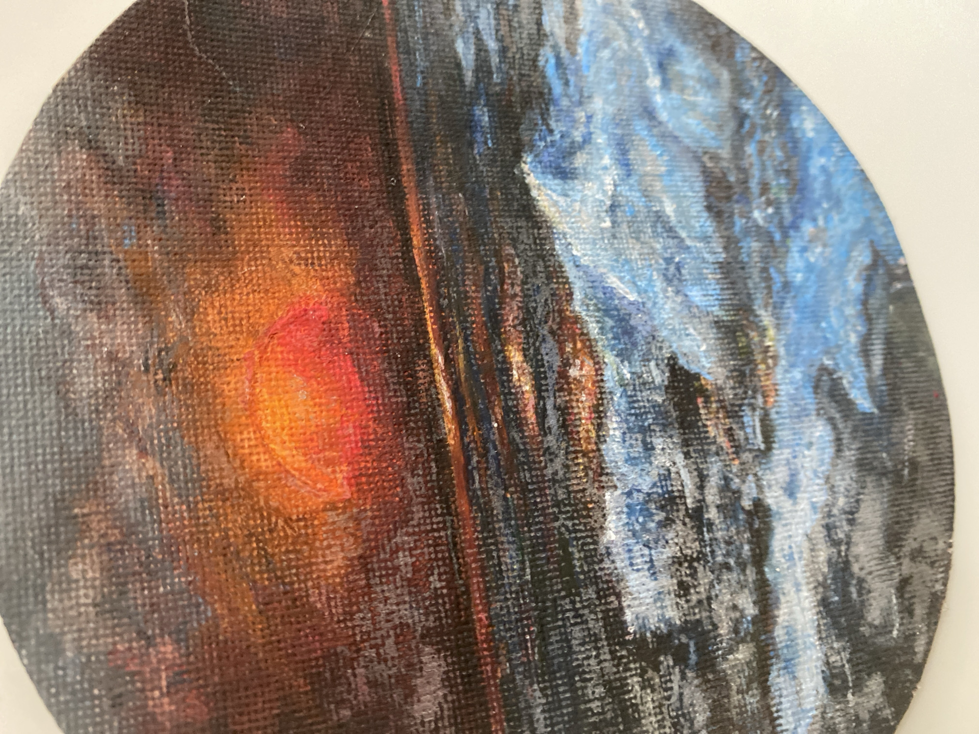 The image is a close-up, circular painting of a sunset over a body of water. The sun is a vibrant orange-red orb low on the horizon, casting a fiery glow on the dark, textured sky filled with deep reds, oranges, and browns. A thin, reflective line of light stretches across the water from the sun. The foreground features choppy waves painted in shades of blue, white, and grey, with highlights suggesting foam and spray. The painting is rendered on a textured canvas. There is no discernible location in Sokhumi, Abkhazia, or any specific details about people, objects, actions, or interactions. The scene is purely naturalistic and abstract.