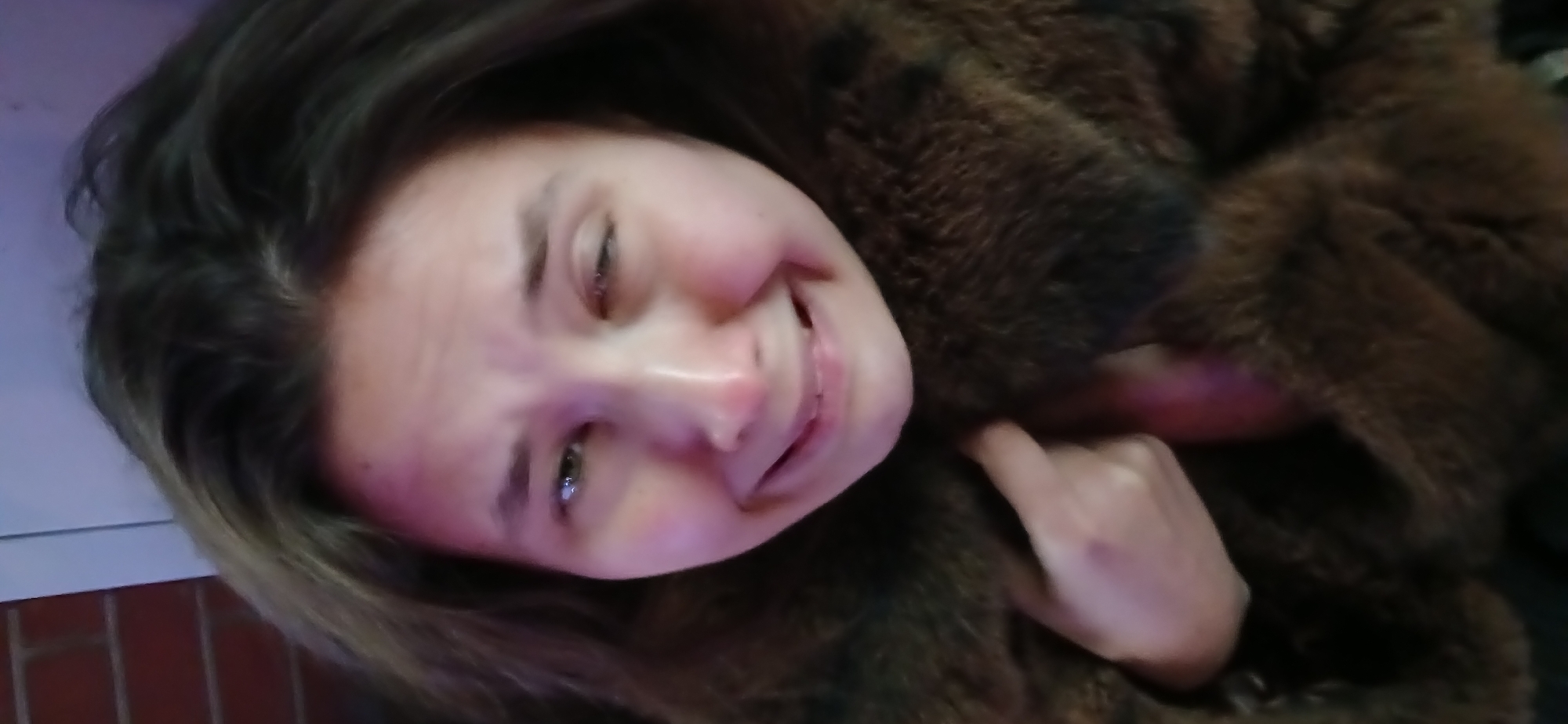 A young woman with long, dark brown hair is captured in a close-up shot, her face dominating the foreground. Her head is tilted, and she is smiling broadly, with her eyes squinted in amusement and her cheeks appearing flushed, conveying a joyful and playful emotion. She is wearing a dark, textured garment, possibly a fur or faux fur coat, which covers much of her body. Her left hand is visible near her face, and her right hand is also partially seen, both appearing to interact with the dark garment or an object within it. A glimpse of a bright pink or purple object is visible near her mouth and also close to her right hand, though its specific nature is indistinct. The background is dark and out of focus, with a small portion of a light-colored surface and a brick-patterned wall visible on the far left, suggesting an indoor or dimly lit outdoor setting. The lighting appears to be artificial. There is no discernible text within the image. The scene is set in Moscow, Russia.