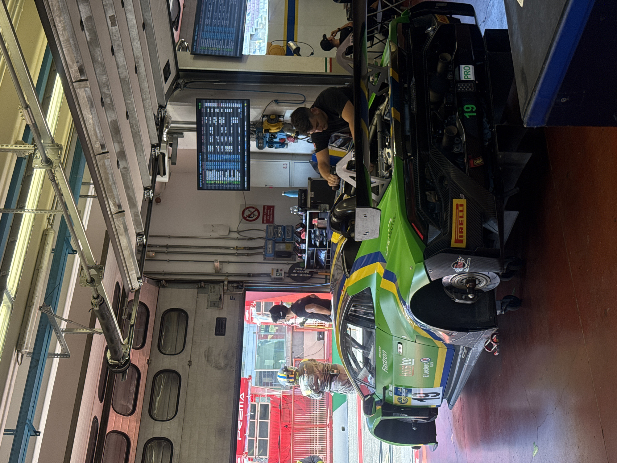 In the pit lane of the Autodromo Internazionale del Mugello in Scarperia, Italy, a green and yellow racing car, identified by the number 19 and "PRO" designation, is positioned on a hydraulic lift with its rear exposed. A mechanic, wearing a black t-shirt, is actively working on the car's engine compartment. Two other individuals, wearing racing suits, stand near the entrance of the garage, observing the scene. One is wearing a helmet with a yellow and blue stripe pattern, and the other is wearing headphones and a dark t-shirt. The garage is equipped with overhead fluorescent lighting and a large roll-up door. Two digital displays hang from the ceiling, showing racing data. In the background, another vehicle, possibly a team transport, is visible through the open garage door. The floor is made of a dark, red-toned material, and there are various pieces of equipment and storage units in the vicinity.