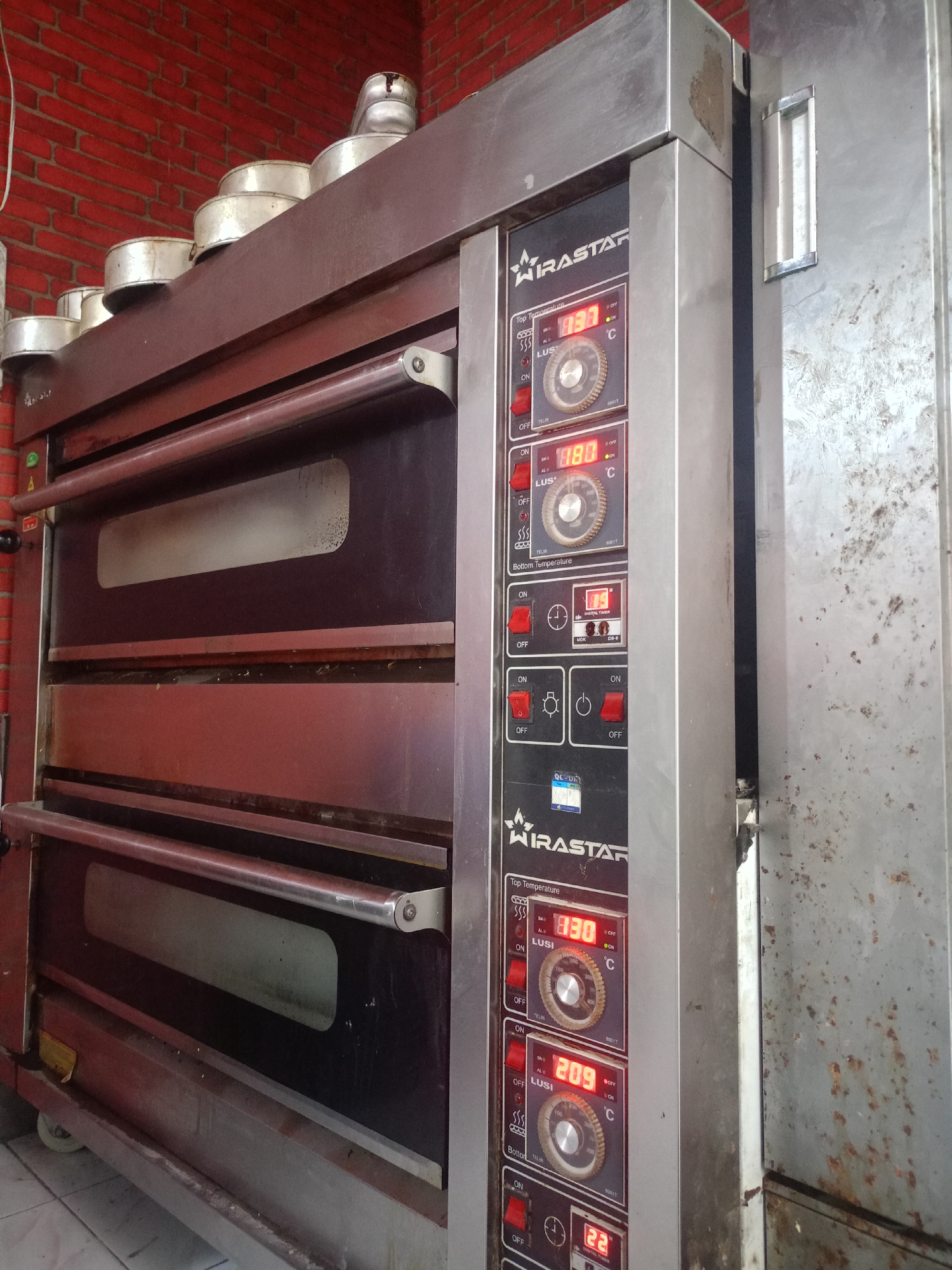 An industrial oven with multiple compartments is shown in a shop in Lumajang, Indonesia. The oven is made of stainless steel and has a dark red front with glass windows. The oven has digital displays that show temperatures and controls for each compartment. The walls of the shop are covered in red brick. On top of the oven are several metal baking pans. The floor is tiled. The oven appears to be in use or ready for use, with digital readouts indicating temperatures of 830, 880, 209, and 22 degrees Celsius.
