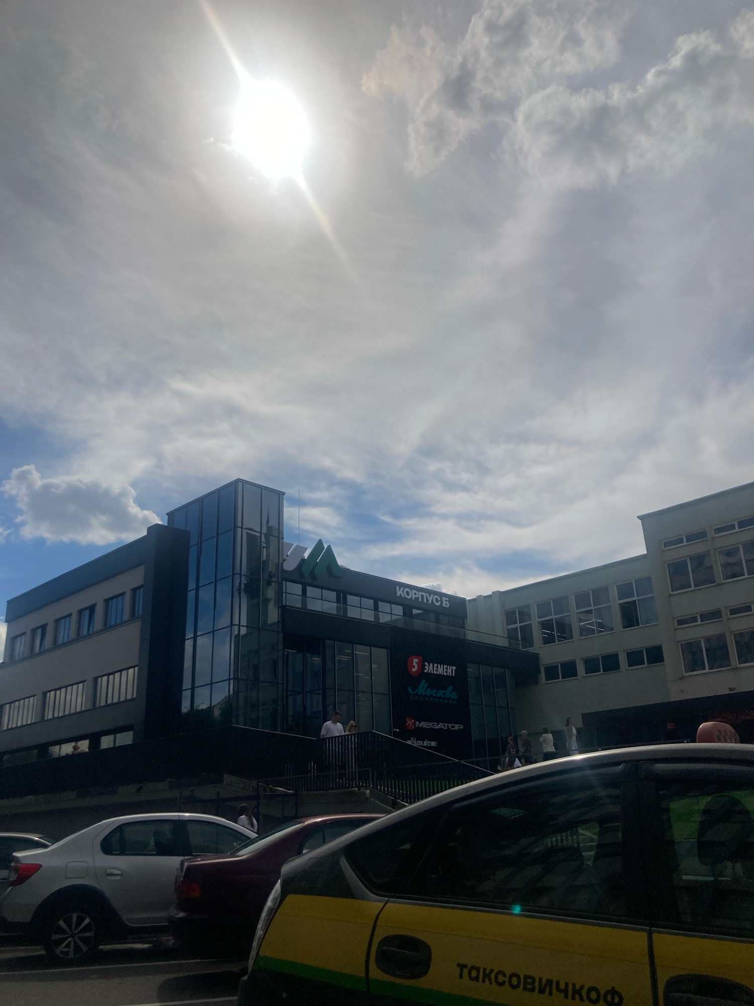 The image shows a modern commercial building complex in the city of Brest, Belarus, under a bright, cloudy sky. The building, labeled "КОРПУС Б" and featuring logos for "5 ЭЛЕМЕНТ" and "MEGATOP", has a prominent glass facade on one section and appears to be a retail or office space. Several cars are parked in front of the building, including a yellow and green taxi with the word "ТАКСОВИЧКОФ" visible on its side. A few individuals are seen standing near the building's entrance and on a walkway. The sun is very bright, causing lens flare in the upper portion of the image.
