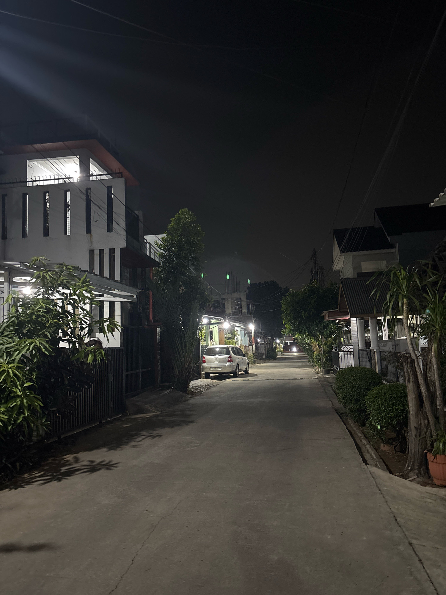 This is a nighttime photograph of a residential street in the city of Bekasi, Indonesia. The street is paved and appears to be a quiet neighborhood. On the left side of the street, there is a modern-looking two-story house with white walls and vertical dark windows. A black metal fence encloses the property, and lush green foliage is visible in front of it. A white car is parked on the street in front of the house.
Further down the street, on both sides, are more houses, some with visible lighting from within. Trees and bushes line the sidewalks and properties, casting shadows on the street. Power lines are visible overhead against the dark night sky. In the distance, the headlights of another vehicle can be seen approaching. The overall impression is one of a calm, suburban evening.