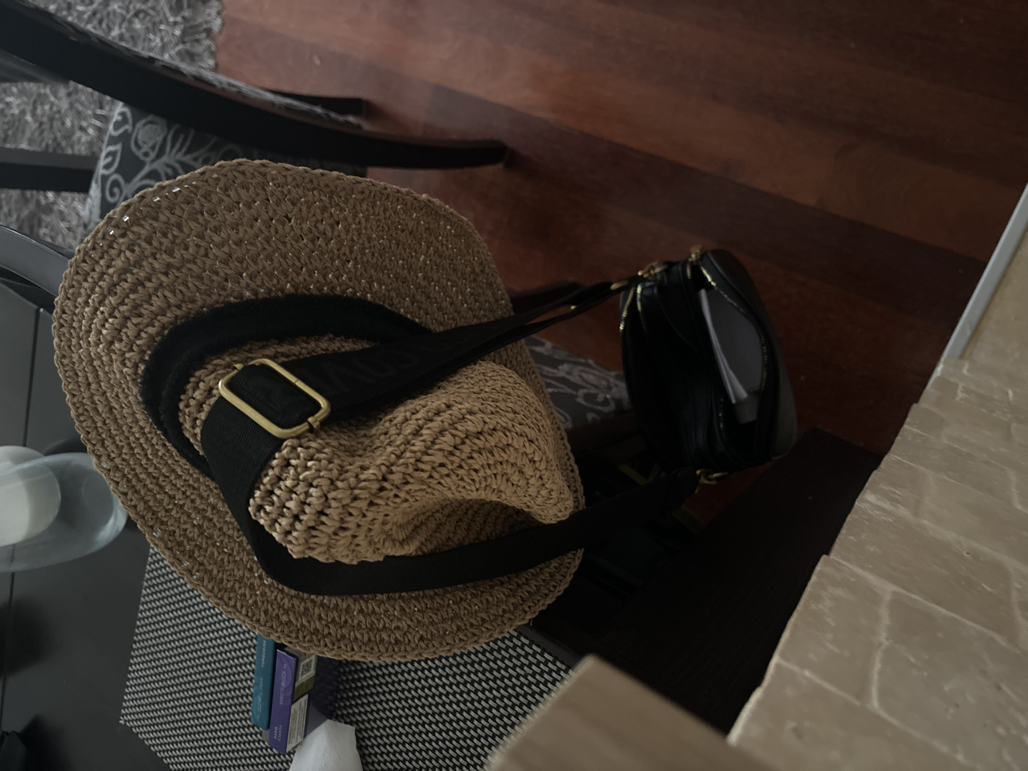 The image shows a tan straw hat with a black band and gold buckle, resting on a black and white patterned surface. Next to the hat is a small, black purse. The scene is set indoors, with a dark wood floor visible in the background and a patterned cushion underneath the hat. To the side, there's a glimpse of a stone-like surface.
There is no verifiable information in the image that confirms it is in Budva, Montenegro.
