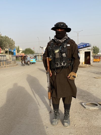 The image shows a man in Kandahar, Afghanistan, standing on a street. He is wearing a brown shalwar kameez, black boots, a black tactical vest, and a black hat. A black scarf covers his face. He is holding a rifle. In the background, there are buildings, vehicles, and people.