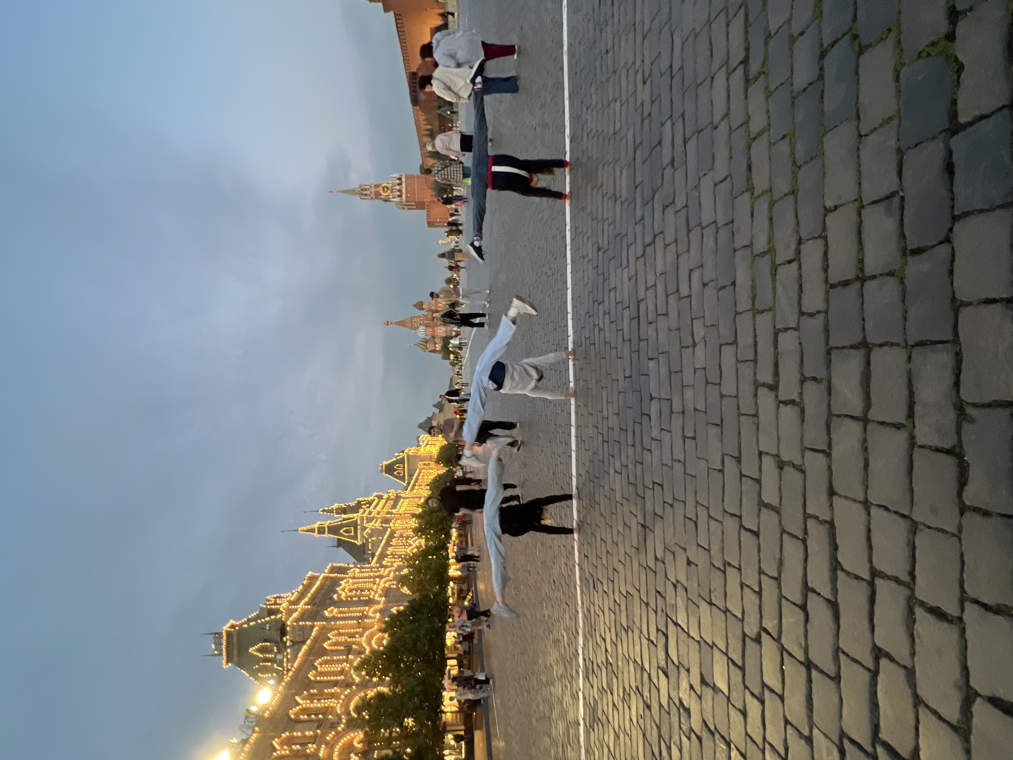 The image depicts Red Square in Moscow, Russia, at dusk. The GUM department store is illuminated with numerous lights on the left, and the Spasskaya Tower of the Kremlin is visible in the background. In the foreground, three individuals are performing cartwheels on the cobblestone pavement. One person is in the center, with their legs extended outwards in a straddle split while inverted, and another individual to the left is in a similar inverted position with legs apart. A third person is also performing a cartwheel further to the right. Several other pedestrians are scattered in the background, observing the scene or walking through the square. The sky is a gradient of blue and gray, indicating the transition from day to night.