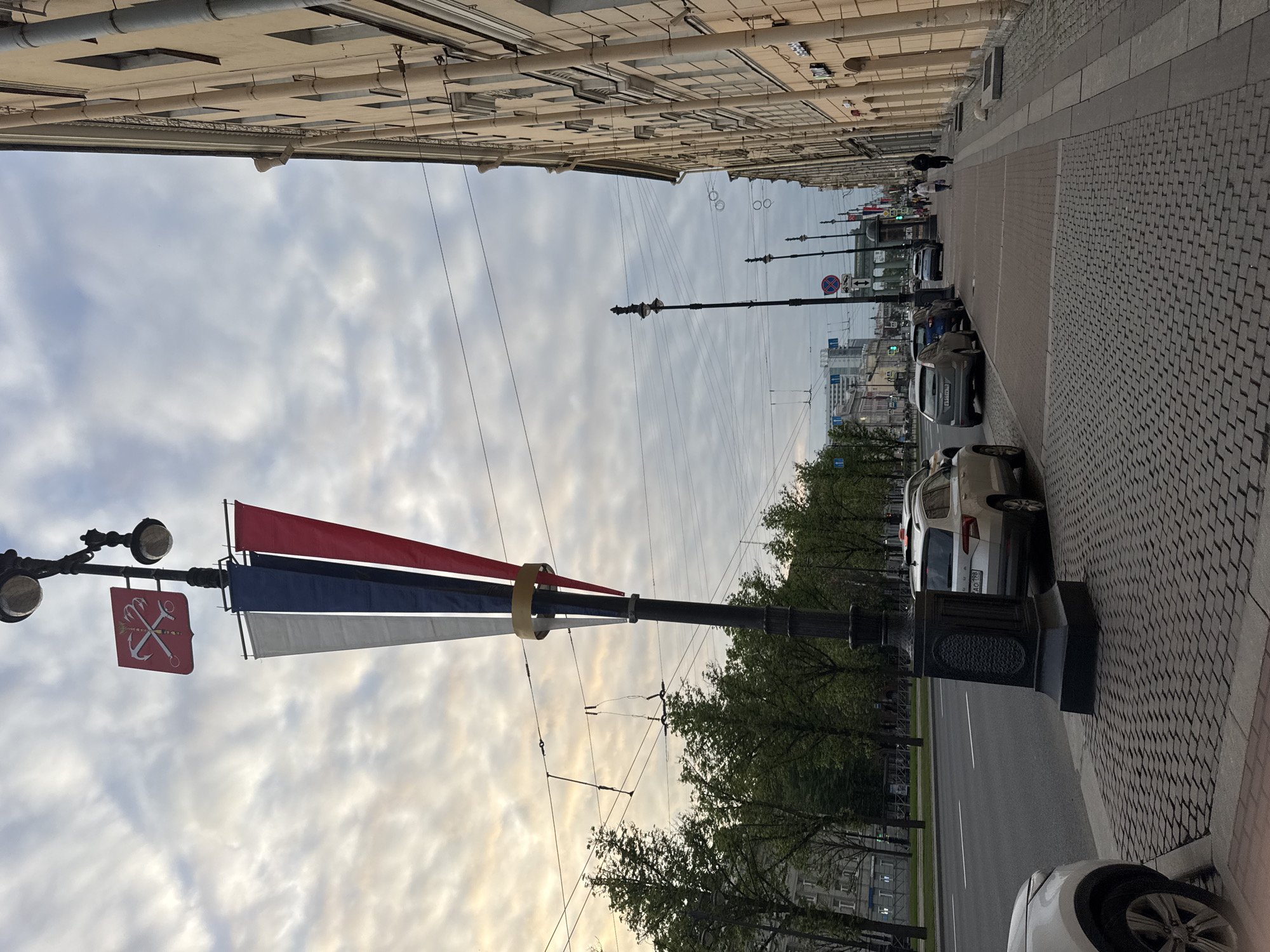 This media file captures an urban street scene in Saint Petersburg, Russia, under an overcast sky. The primary perspective is along a wide, grey brick-paved sidewalk, extending into the distance.
In the immediate foreground, a black, ornate lamppost stands, adorned with three rectangular banners in red, blue, and white, resembling the Russian national colors. Below these banners, a red square shield hangs, featuring a silver anchor crossed with a pickaxe and shovel—a prominent emblem associated with Saint Petersburg.
Parallel to the sidewalk on the left is a paved street, where several cars are parked. One silver SUV displays a partially visible Russian license plate with "AO 198", indicating the Saint Petersburg region. Further down the street, additional lampposts, power lines (possibly for trams), and other vehicles are visible, along with a red circular "no stopping" traffic sign. A line of green trees separates the street from the buildings on the far side.
In the mid-ground, a few indistinct figures of people can be seen walking along the sidewalk. The background is dominated by a row of multi-story buildings featuring classical architectural elements such as columns and decorative facades, characteristic of Saint Petersburg's urban landscape. The sky is largely covered by white and grey clouds, suggesting an overcast day with soft, diffused light.