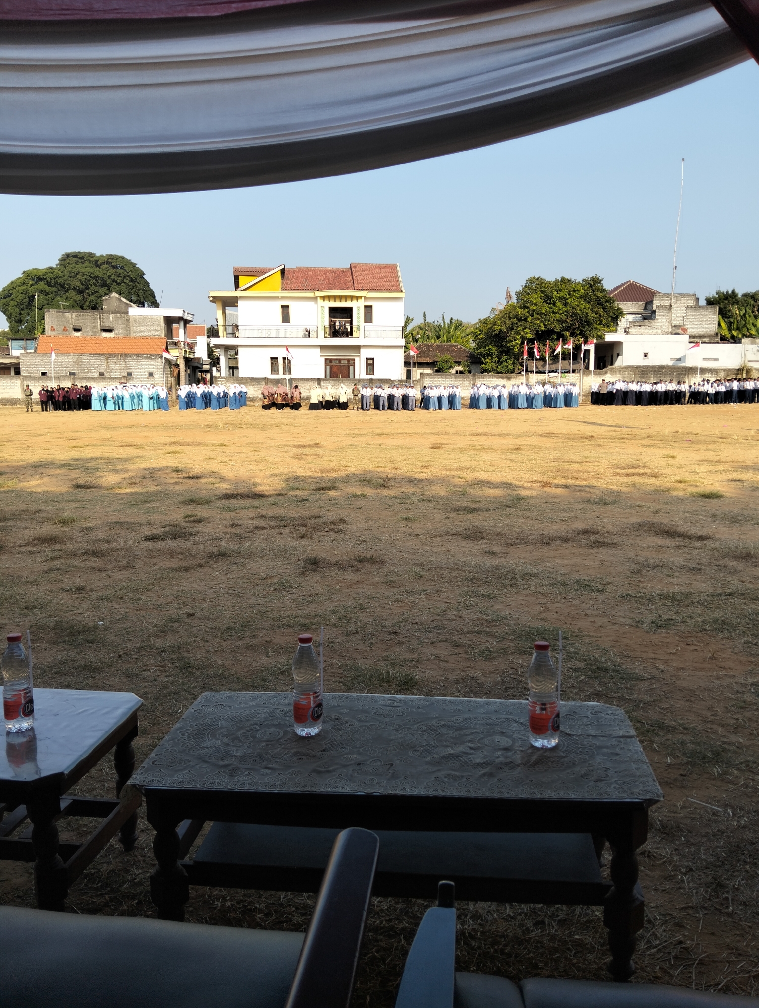 The image depicts an outdoor gathering in Paciran, Indonesia, possibly a school event or ceremony. In the foreground, two dark-colored tables are set up, each with a clear plastic bottle of water. The ground is covered in dry grass and dirt, indicating a field or open area. In the middle ground, a line of people, many wearing white and blue uniforms, stand in formation. Behind them, several buildings are visible, including a white building with a yellow roof and a taller brick structure. Flags are planted in front of the buildings. The sky is clear and blue. The overall impression is one of an organized event taking place in a sunny, open environment.