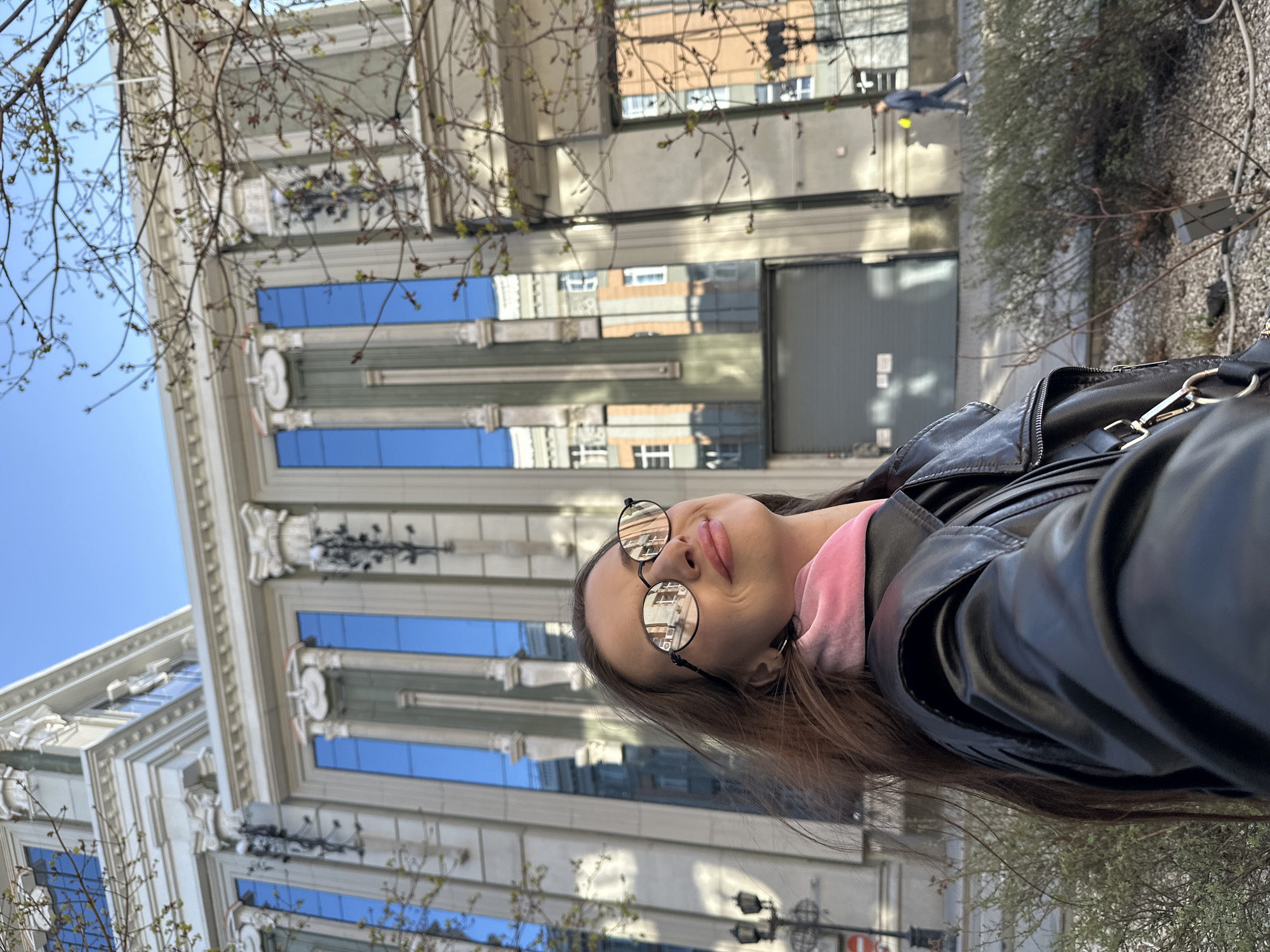 The image depicts a selfie of a woman with long brown hair, wearing a black leather jacket, and round sunglasses with reflections of buildings. She is also wearing a pink garment underneath the jacket.
The backdrop reveals an architectural building with ornate detailing, columns, and reflective blue windows, suggesting a governmental or historical building. This building is the Government Offices building located in Yekaterinburg, Russia. The architecture and the reflection in the sunglasses match that structure.
The sky is blue with no clouds visible, indicating clear weather. Some bare tree branches are in the foreground near the building. A man in a suit is walking in the background. Additionally, a bush is on the right side of the woman.