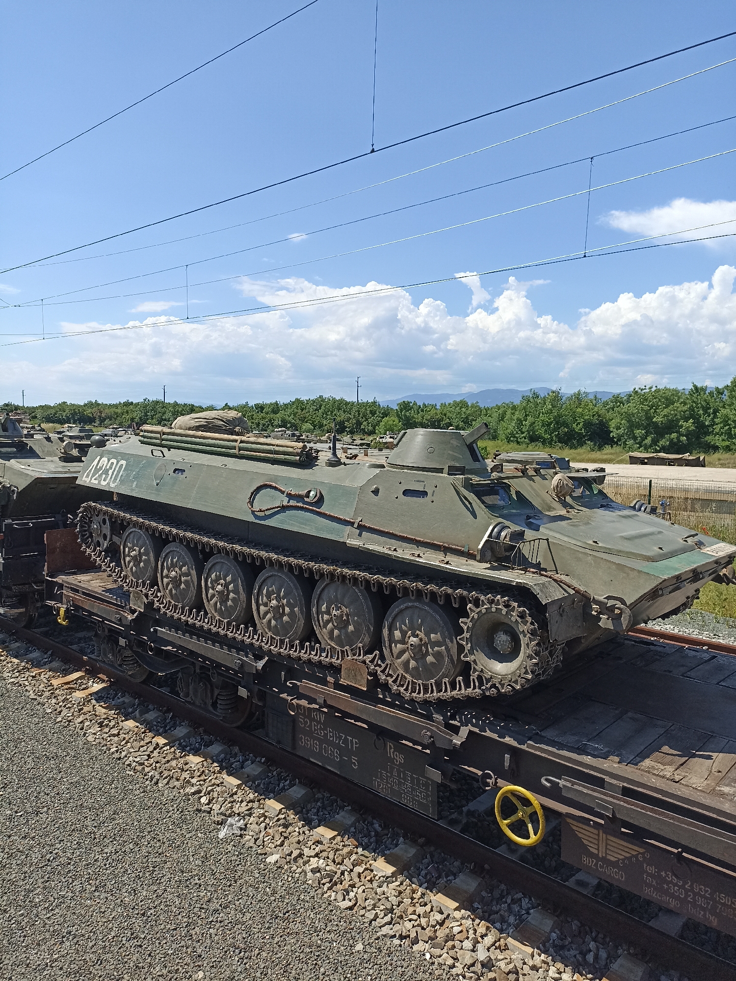 A green, tracked, armored personnel carrier (APC) is secured on a flatbed railcar. The APC has the number "4230" painted on its side. The flatbed railcar has markings and information printed on it, including "3T RIV," "52 BG-BDZ TP," "3919 066-5," "Rgs," "TAIBICI 15140-148-156," "120t - 009," "BDZ CARGO," and contact information for BDZ Cargo. The railcar also features a bright yellow wheel-shaped mechanism on its side. The image is taken outdoors on a sunny day with a blue sky and scattered white clouds. In the background, there is a tree line and possibly some low mountains in the distance. Gravel surrounds the railroad tracks. While the location is suggested to be Zimnitsa, Bulgaria, there are no identifying features in the image itself to confirm this.