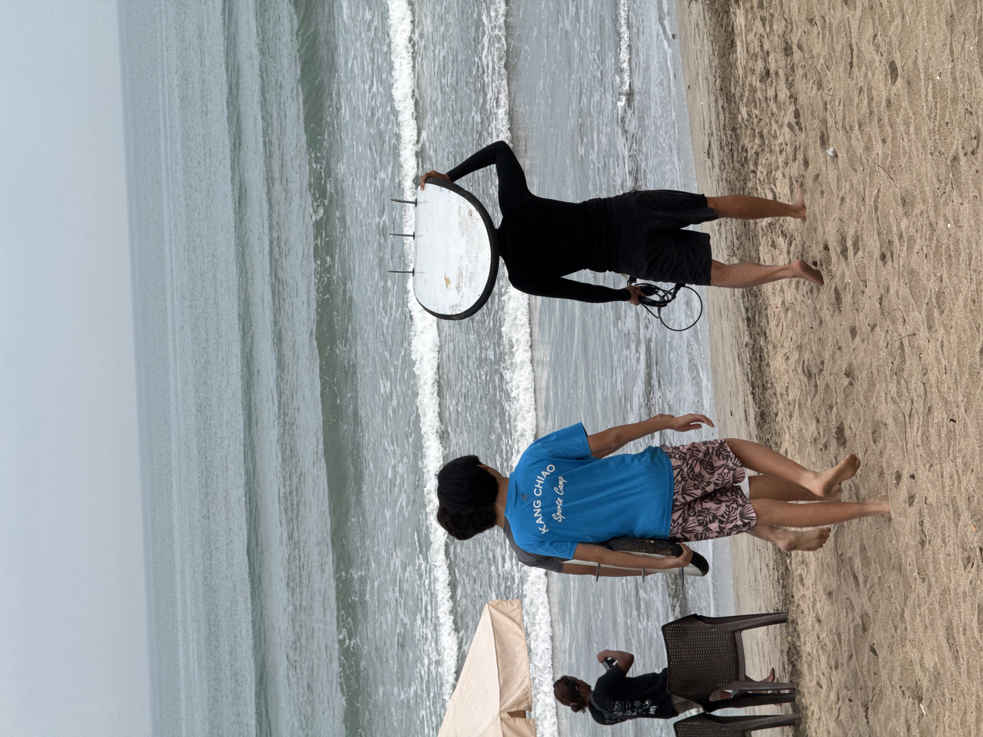 Three individuals are walking on a sandy beach in Kuta, Indonesia, towards the ocean. The person in the foreground, on the right, is wearing a black long-sleeved rash guard and black shorts, carrying a surfboard on their head and a coiled leash in their left hand. They are facing away from the camera, towards the water. To their left, two other individuals are walking together. The one on the right is wearing a blue t-shirt with the words "KANG CHIAO Sports Camp" and patterned shorts. The person to their left is wearing a dark grey t-shirt and is partially obscured. To the far left of the frame, a person is standing and appears to be taking a photograph with a phone, with a beach umbrella and chairs visible nearby. The ocean is visible in the background, with waves rolling in under a cloudy sky. The sand is light brown and textured.