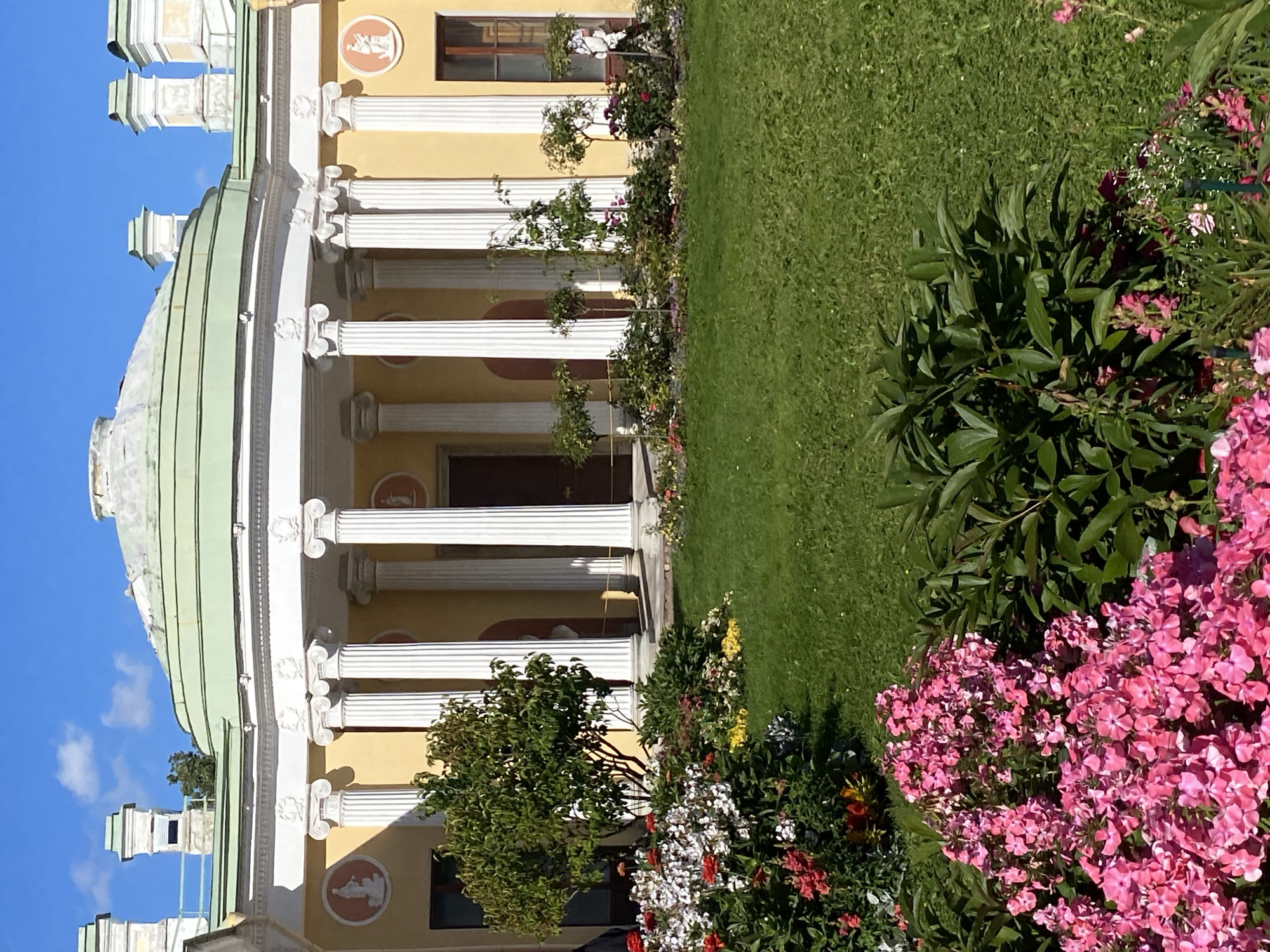 The image captures a sunny day in the city of Pushkin, Russia, showcasing a classical architectural facade adjacent to a lush garden. The building features a semicircular portico supported by tall, fluted white columns. The walls of the building are painted a warm yellow, and decorative circular medallions with reliefs are visible above some of the windows. A pale green dome caps the central section of the building. The garden in the foreground is meticulously maintained, with a vibrant green lawn and a variety of flowering plants in shades of pink, white, and red. Small trees and shrubs are strategically placed throughout the garden, adding depth and texture to the scene. The sky above is a clear, bright blue with a few wispy clouds. The overall impression is one of serene beauty and historical grandeur.