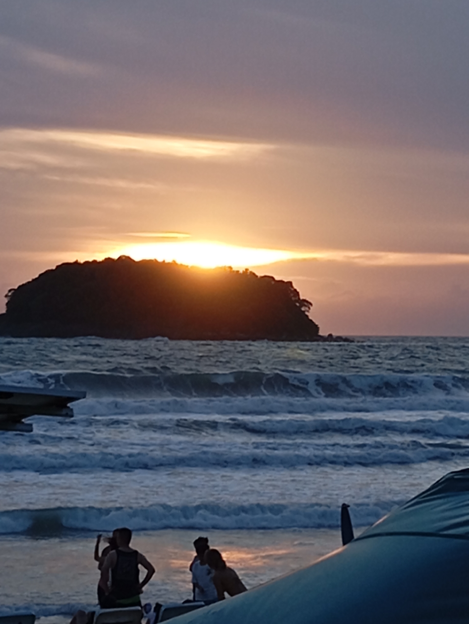The image depicts a sunset over the ocean in Karon, Thailand, with a silhouetted island in the distance. The sky is a gradient of purple and orange, with a bright sun peeking from behind the island. Waves crash on the shore, and several people are visible in the foreground, sitting on chairs or standing near the water. A large blue structure, possibly a beach umbrella or canopy, occupies the lower right portion of the frame. The scene conveys a peaceful, tropical atmosphere.