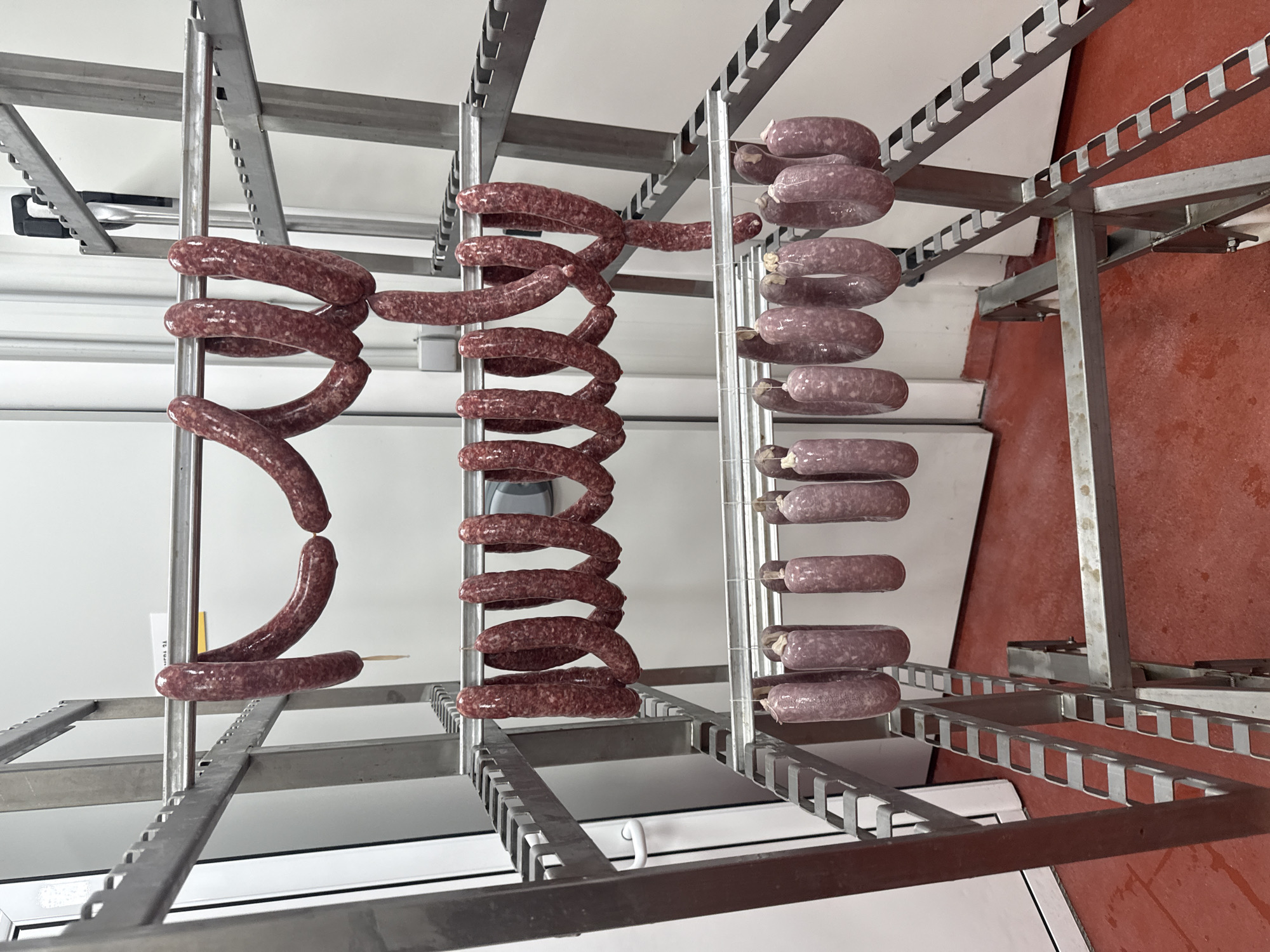 The image shows rows of sausages hanging on metal racks in a room with mixed white and red flooring. Three rows of sausages are visible: one row of single sausages pierced by skewers and hanging vertically, and two rows of coiled sausages. The metal racks have a tiered, slotted design. The room has a clinical appearance, suggesting a production or storage facility. The location in the city of Stara Zagora, Bulgaria, is not apparent from the visual elements.
