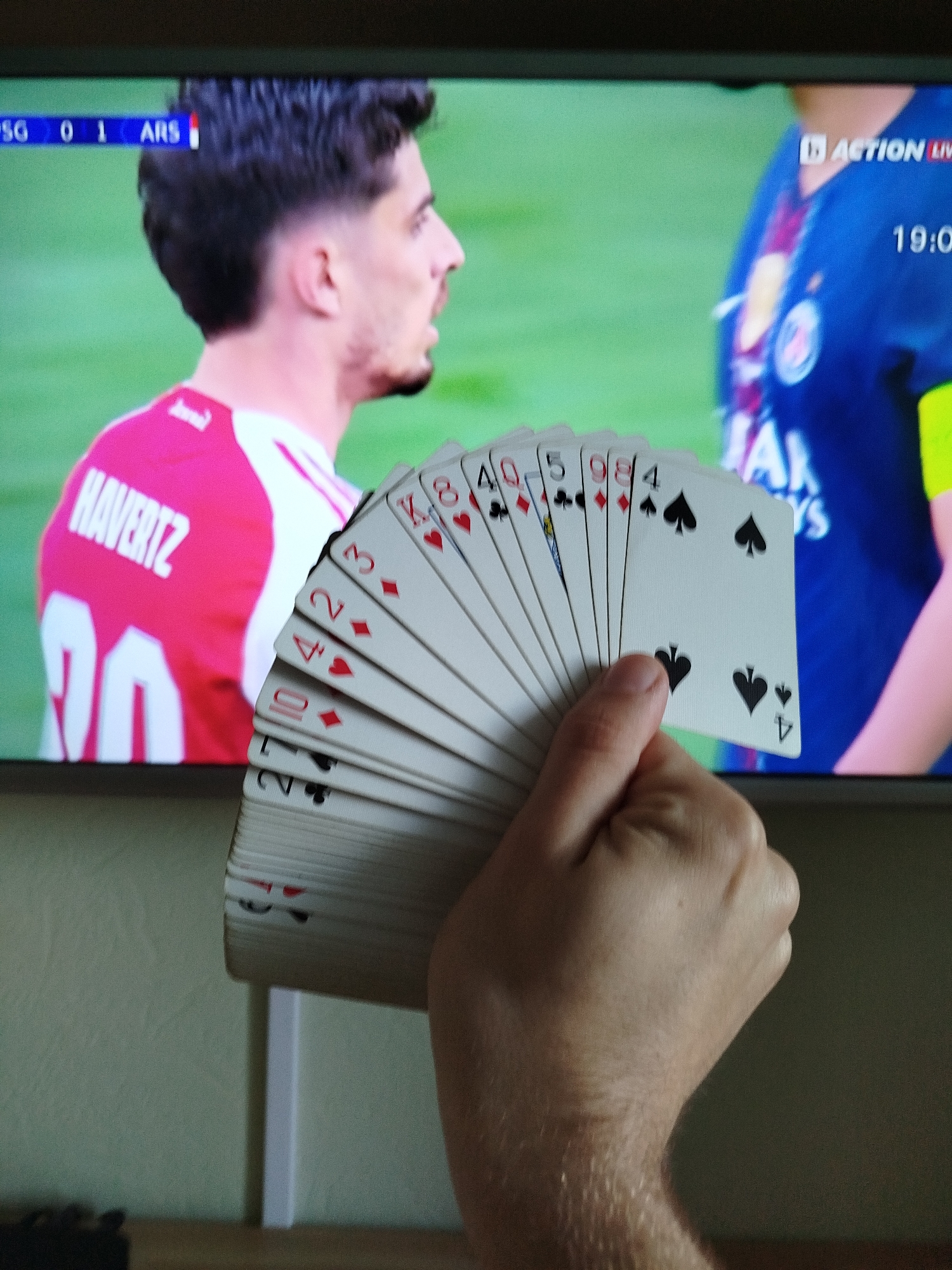 A person is holding a hand of playing cards fanned out in front of a television screen. The television displays a soccer match, with "PSG 0 1 ARS" visible at the top left and "HAVERTZ" with a number "29" on the back of a red jersey. The soccer match's time is "19:05". The playing cards visible are, in order, 4 of Spades, 9 of Diamonds, 5 of Clubs, 10 of Diamonds, 4 of Hearts, 2 of Diamonds, and King of Diamonds. The person's hand is fair-skinned and slightly hairy. The background behind the hand and television is a light green/beige wall. There is no information about the location of the image being in Varna, Bulgaria.