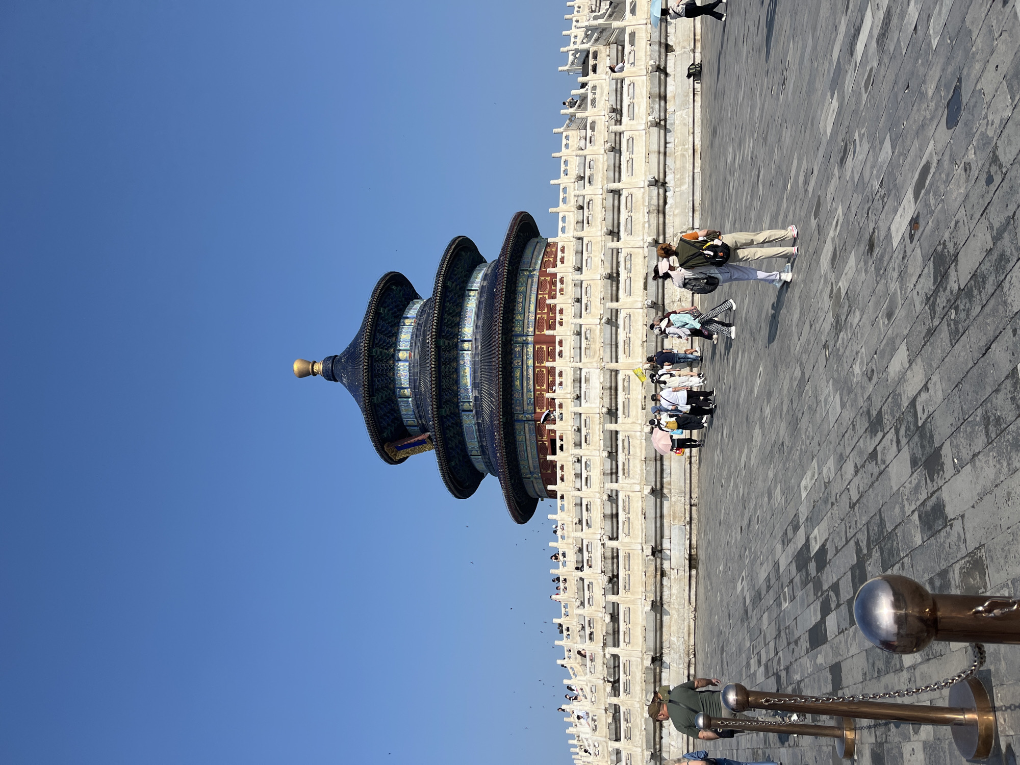 The image shows the Hall of Prayer for Good Harvests, a key structure within the Temple of Heaven complex in Beijing, China. The building is a tripartite circular structure with a blue tiled roof topped by a golden finial. It stands on a tiered white marble terrace surrounded by a balustrade. Numerous people are present on the terrace and in the foreground, many appearing to be tourists. Some are walking, while others are standing and observing the architecture. The ground in the foreground is paved with large grey stone slabs, some of which are wet, indicating recent rain or cleaning. The sky is a clear, bright blue. The overall scene depicts a sunny day at a historical and cultural landmark.