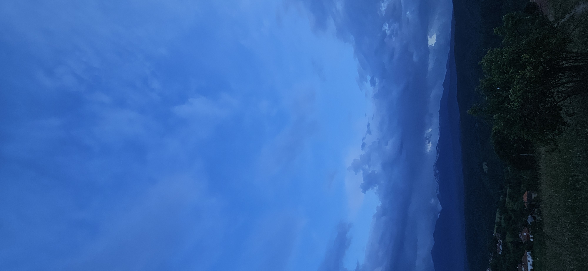 The image presents a dramatic landscape scene, likely captured at dusk or late evening, with a prominent, cloudy sky dominating the view. The sky transitions from a lighter, deep blue on the left to darker, more ominous grey-blue clouds on the right, suggesting a build-up of weather or a recent storm.
Below the heavy clouds, a dark mountain range stretches across the midground from right to left, its peaks silhouetted against the sky. Hints of lighter patches, possibly snow or cloud formations, are visible on some of the higher ridges. On the far right and extending into the foreground, dense, dark green trees and foliage are visible, indicating a forested hillside.
At the bottom right of the image, nestled into the landscape, a small village or settlement is discernible, with several houses featuring reddish-brown roofs and lighter walls. A patch of dark grass occupies the very bottom right foreground. The overall lighting is dim and muted, contributing to a tranquil yet somewhat dramatic atmosphere. There is no visible text in the image. The scene is consistent with the mountainous and rural environment of Gutsal, Bulgaria.