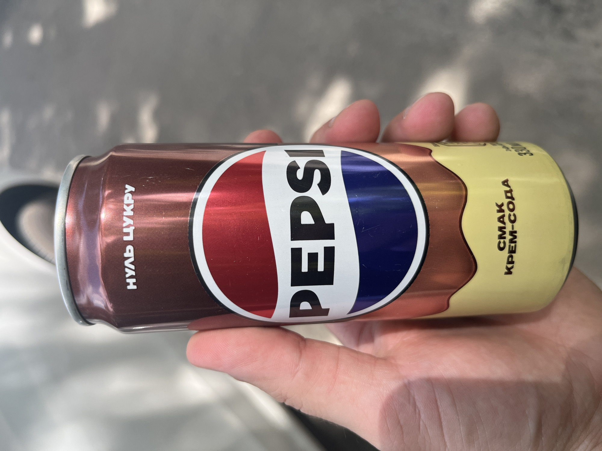 A hand holds a Pepsi can horizontally. The can has a brown, metallic design with a wave of yellow liquid flowing down from the top on the right side. The Pepsi logo, a red, white, and blue circle with the word "PEPSI" in white text, is prominent on the can.
To the left of the Pepsi logo, text in white Cyrillic letters is visible. It appears to read "НУЛЬ ЦУКРУ", which translates to "ZERO SUGAR". To the right of the Pepsi logo, more white Cyrillic text is visible, reading "СМАК КРЕМ-СОДА" which translates to "CREAM SODA FLAVOR". The volume "330" is also visible.
The background is a blurred outdoor scene with sunlight casting shadows, suggesting it is daytime. The setting appears to be an urban environment, possibly a street or sidewalk, given the concrete-like texture and lighting. The image conveys a casual, everyday moment of someone holding a beverage. There are no strong emotional cues, but the image feels neutral and observational. The location context is Kharkiv, Ukraine.
