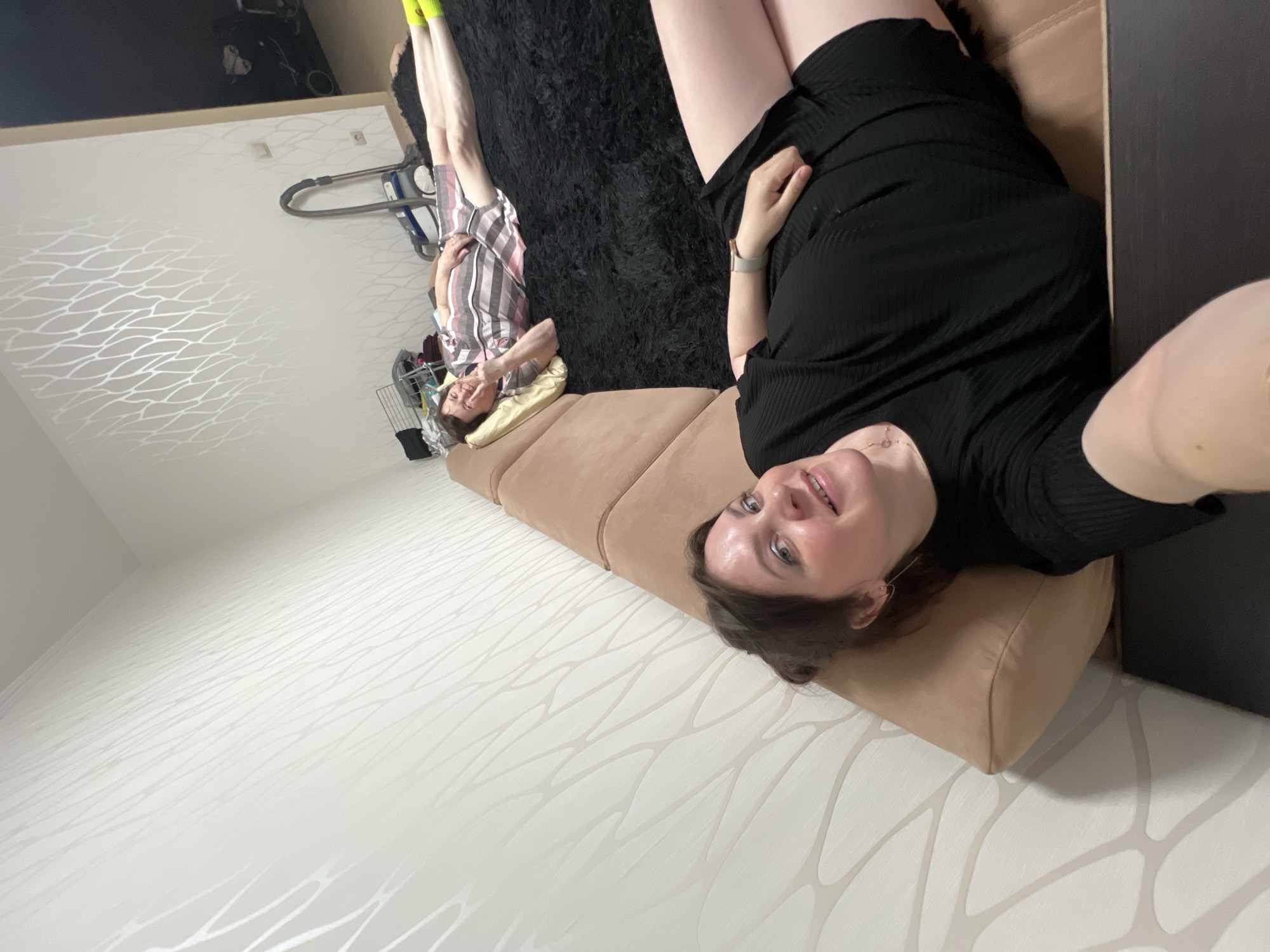 This media file captures an indoor scene, likely a residential living space or bedroom, featuring two women relaxing. The image is rotated, but when viewed upright, the perspective is from the vantage point of one person taking a selfie or being photographed while lying down.
The person closest to the camera is a young to middle-aged woman, lying on a light brown or beige sofa. She has dark hair, a light complexion, and is looking up towards the camera with a gentle smile. She is wearing a black, ribbed short-sleeved top or dress, and a delicate necklace is visible around her neck. A watch or wristband is on her left wrist, and parts of her legs are visible.
Further into the background, another woman, appearing older, is seen lying on a dark, shaggy rug. She is also smiling and making a "peace" or "V" sign with her hand. She has short, dark hair, wears glasses, a striped shirt (featuring pink, white, and darker stripes), and bright yellow socks. Her head rests on a light yellow pillow, and her legs are extended.
The room features light-colored walls with a distinctive, subtle pattern of wavy, intertwined lines. In the background, near the further woman, a grey and blue upright vacuum cleaner is visible against the wall. A metal wire structure, possibly a small laundry basket or part of a drying rack with some clothes, is also seen nearby. Electrical outlets are present on the far wall, and a dark horizontal strip, possibly a mounted television or shelf, runs along the upper part of the wall.
Both individuals appear relaxed and happy, suggesting a casual moment of leisure. The lighting suggests daytime or a well-lit interior, with no specific outdoor or time-of-day cues. The setting is a comfortable and lived-in residential space. No visible text is present in the image.