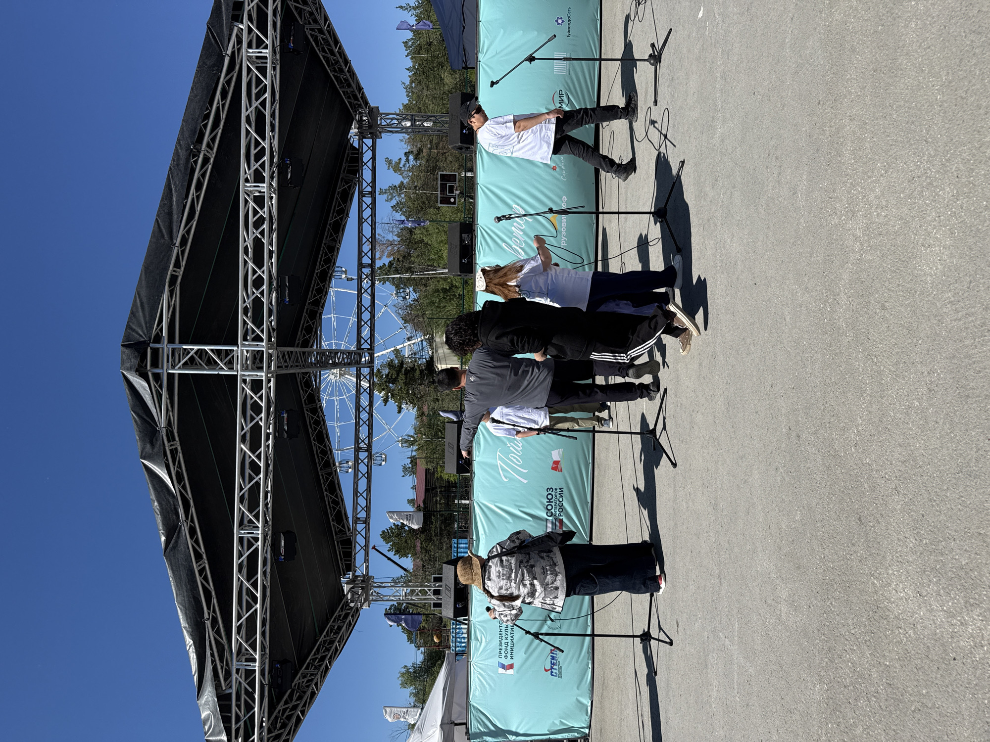 A group of people are gathered on an outdoor stage, possibly for a performance or event. The stage structure is made of metal trusses and is covered with a black tarp. Several microphones are set up on stands in front of the stage. The backdrop behind the performers consists of several light blue banners with text in Russian. One banner reads "СОЮЗ ЛИТЕРАТОРОВ РОССИИ," which translates to "UNION OF WRITERS OF RUSSIA." Another banner has the word "МИР" (Peace) visible.
The people appear to be standing and interacting. One individual in a white shirt and black pants and cap is standing closest to the microphones, perhaps singing or speaking. Others are standing behind them, some with their backs to the camera. The scene suggests a sunny day, with clear blue skies and strong shadows cast by the people and stage equipment, indicating it is likely midday. In the background, trees and a Ferris wheel are visible, suggesting the event is taking place in a park or recreational area. The overall atmosphere appears to be casual and public. The provided text also includes "ПРЕЗИДЕНТСКИЙ ФОНД КУЛЬТУРНЫХ ИНИЦИАТИВ" which translates to "Presidential Fund for Cultural Initiatives."