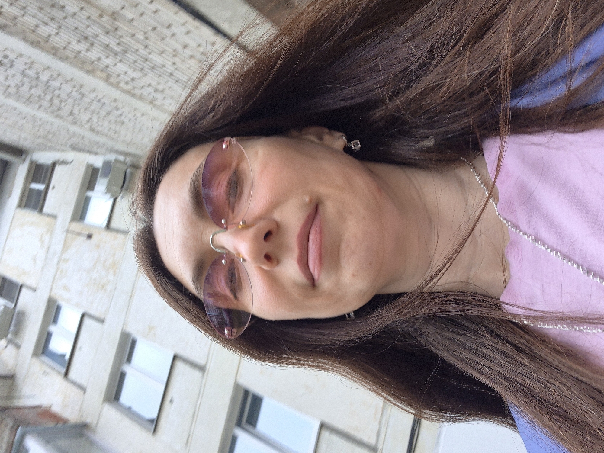 A close-up, rotated portrait-style image features a person with long, flowing dark brown hair, captured outdoors during daylight. The subject is wearing rimless sunglasses with light pink-tinted lenses and a subtle metal frame across the top and nose bridge. A delicate silver chain necklace is draped over a light pink top, and a small, square silver earring is visible in their right ear (viewer's left). The person's expression is serene, with a soft, relaxed smile and closed lips. A small mole is visible on their cheek.
The background shows a light-colored, multi-story building, likely an apartment block or office building, featuring a textured facade and several rectangular windows. Reflections in the windows suggest clear skies. The photograph is significantly rotated clockwise, causing the building to appear at a steep diagonal angle and the subject's head to be tilted towards the upper left. The lighting indicates it is daytime. While the architecture is generic, the provided context situates the location in Chaikovsky, Russia. No text is visible in the image.