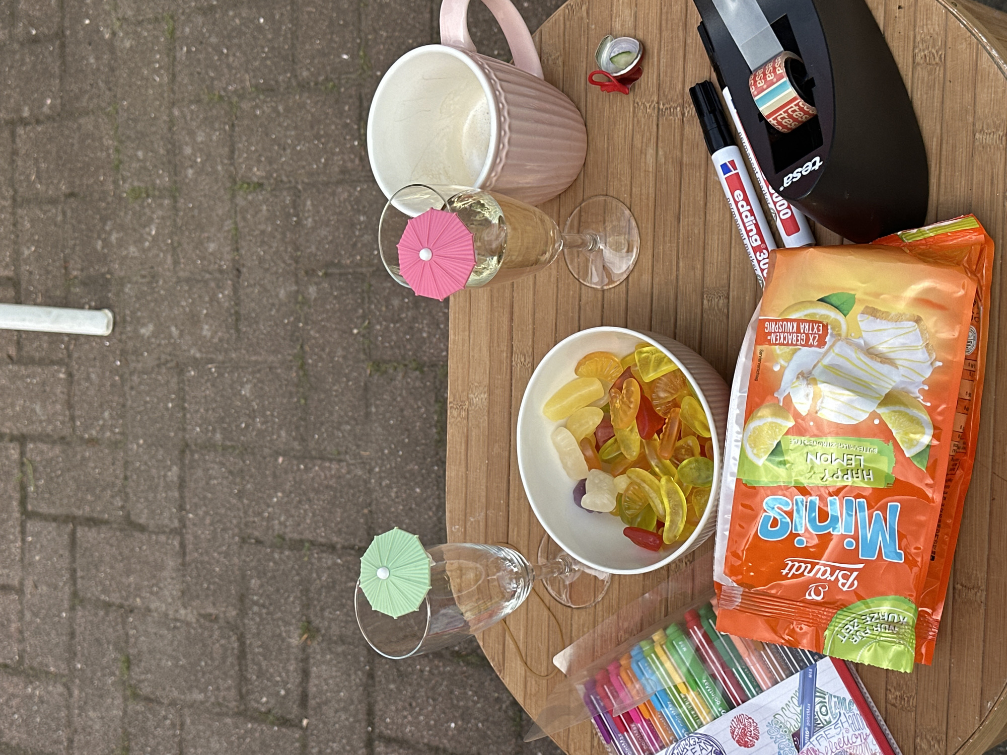 The image depicts a tabletop scene, likely outdoors given the view of pavement in the background. On the table, there is a pink mug, two wine glasses (one containing a liquid and adorned with a pink paper umbrella, the other with a green paper umbrella), a white bowl filled with gummy candies, a package of "Minis" lemon-flavored cookies, markers, a roll of "tesa" tape in a dispenser, and a small container. The "Minis" package is brand name "Brandt" and includes the description "2X GEBACKEN- EXTRA KNUSPRIG" and "BUTTERMILCH ZITRONESTYLE." The markers are identified as "edding 3000 permanent marker." The small container appears to have food contents with a garnish. The scene suggests a casual snack or activity setup. The city and country are mentioned in the prompt, Bargstedt, Germany, though these are not overtly discernible within the image itself.
