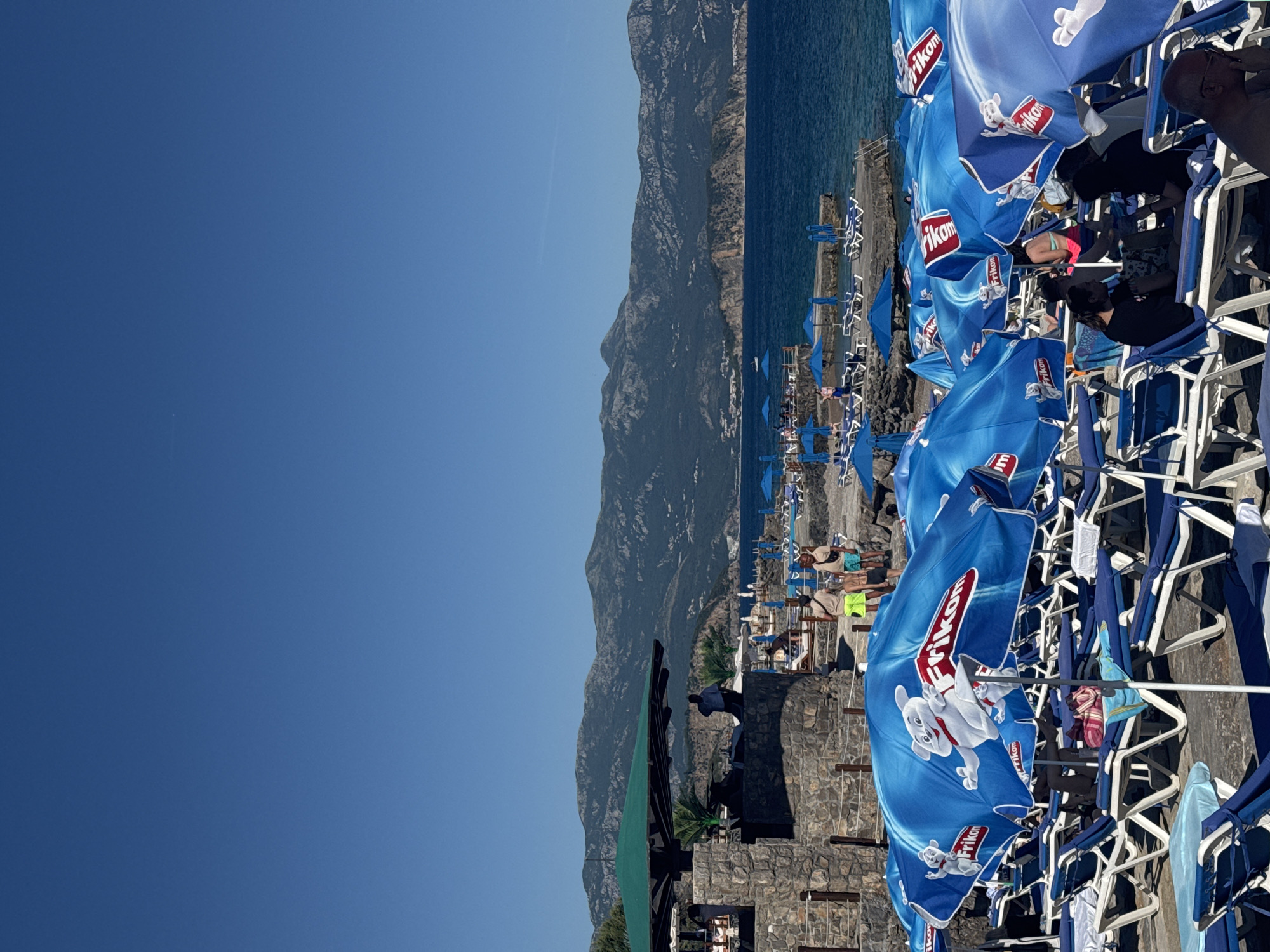The image depicts a beach scene at Prijevor, Montenegro. The foreground is filled with rows of beach umbrellas, mostly blue with the 'Frikom' logo, and white beach chairs. People are seated on the chairs under the umbrellas, though the image does not clearly show individuals' features. Beyond the beach area, the Adriatic Sea extends toward a large mountain range in the background. The sky is clear and blue. A stone building is visible toward the left of the beach area, with a green awning partially covering it.
