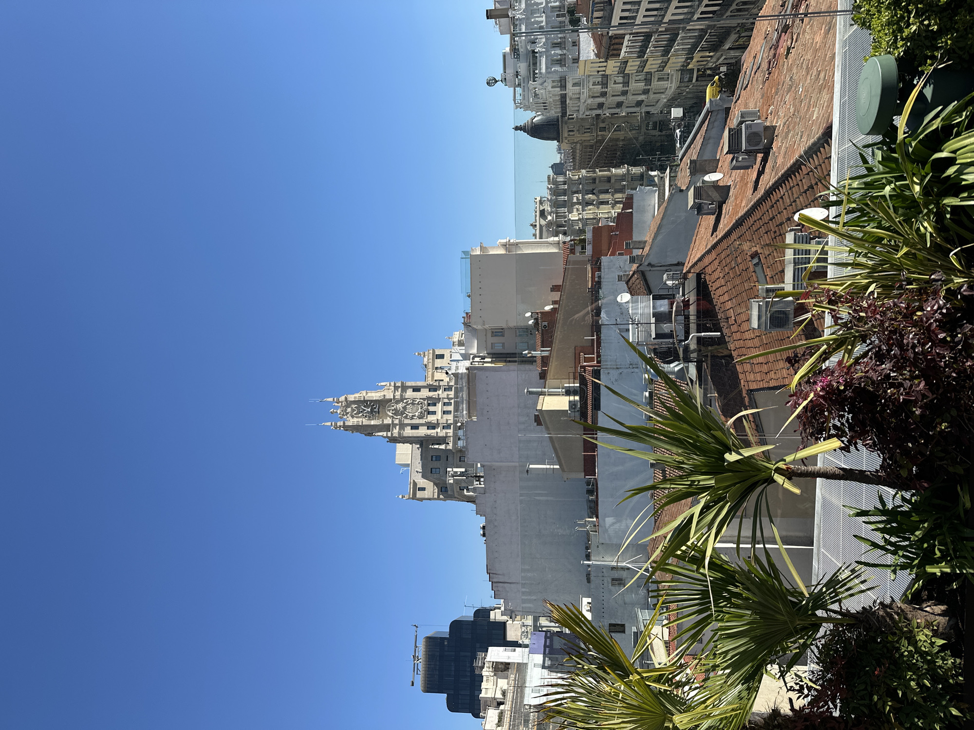 An outdoor, angled medium-shot reveals a cityscape in Madrid, Spain, under a clear blue sky. Predominantly, a mixture of residential and commercial buildings define the urban landscape. Rooftops are visible, marked by diverse architectural styles, from aged terracotta tiles to contemporary flat surfaces, speckled with air conditioning units and other mechanical hardware. Plant life, including palm trees and shrubbery, is present on a rooftop or balcony, offering a contrast to the urban surroundings. The iconic Metropolis Building stands out, showcasing its intricate facade and clock tower.