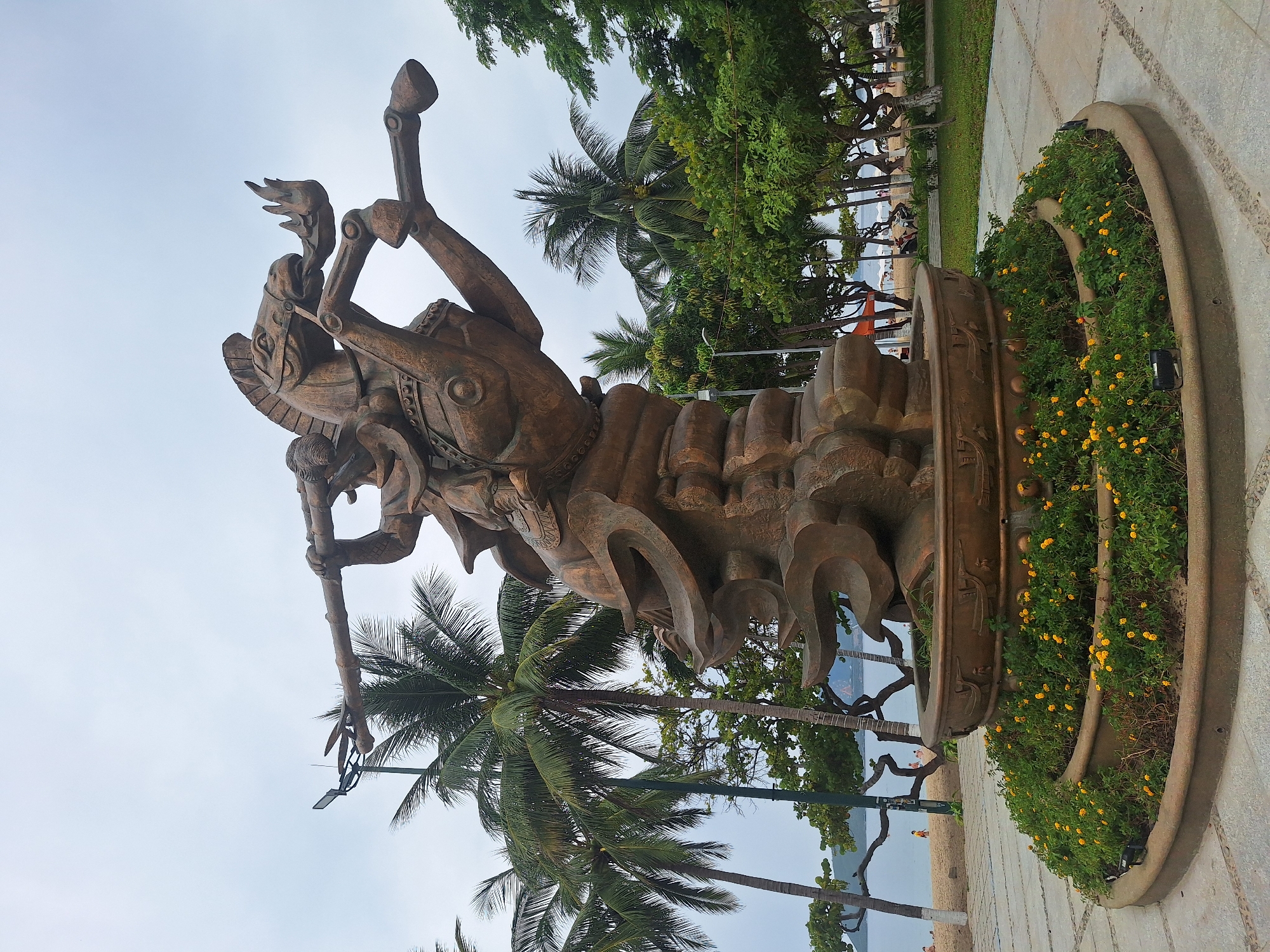 The image shows a large, bronze-colored sculpture of a warrior on horseback in a dynamic pose. The warrior is holding a long spear or staff horizontally. The horse is rearing up on its hind legs. The sculpture is mounted on a tiered, circular base, which is surrounded by a planter filled with green foliage and small yellow flowers. The sculpture and planter are situated in a paved area, likely a public plaza or park.
In the background, there are numerous palm trees and other lush green trees. Beyond the trees, a body of water, likely the sea, is visible, with a beach and a few people on it. The sky is overcast and gray. The overall impression is of a coastal city environment. This sculpture is located at the entrance to the VinWonders amusement park on Hon Tre Island, Nha Trang, Vietnam, and is often referred to as the "Dragon Statue" or the "Statue of the Warrior on Horseback."