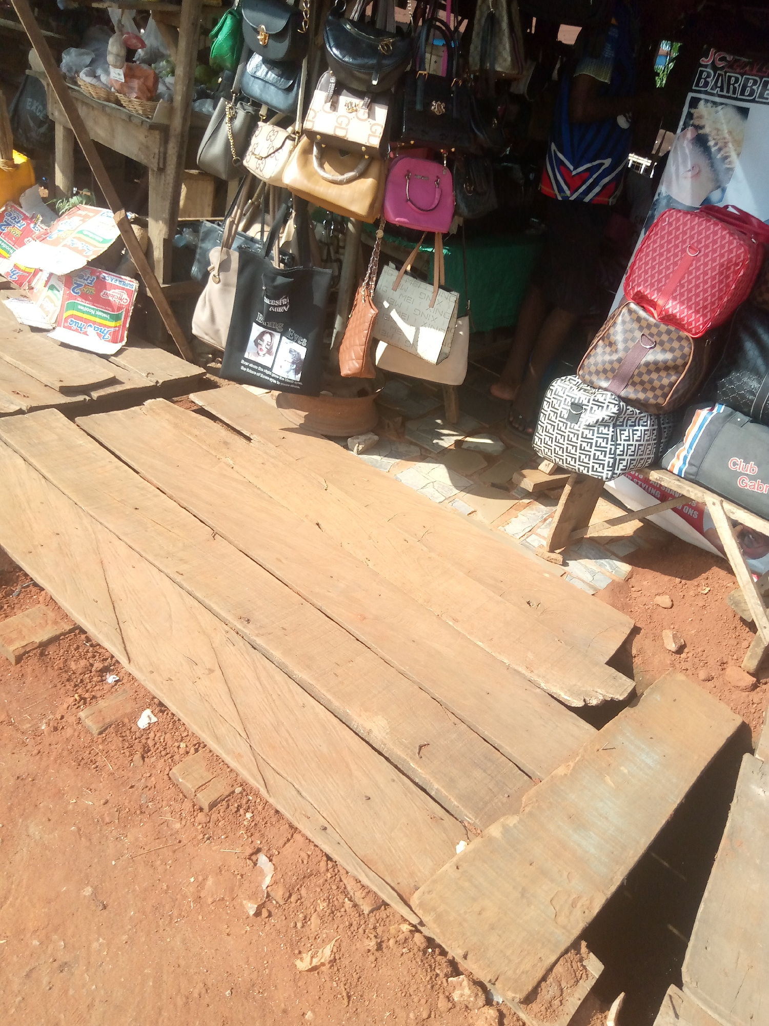This media file shows a street market stall in Nsugbe, Nigeria, laden with various handbags. The stall appears to be a makeshift structure with wooden planks forming the display area and roof. Numerous handbags are displayed, varying in color, size, and design. Some are brightly colored, like a pink handbag, while others are more subdued, such as black and brown leather bags. Several bags feature prominent brand logos or patterns.
On the left side of the stall, stacks of what appear to be instant noodle packets are visible. The ground is covered in reddish soil, characteristic of the region. In the background, a person, partially obscured, is standing behind the stall, presumably the vendor. A poster with text including "JCALE BARBE" is attached to the stall's structure.
The overall impression is a busy market scene, likely in the daytime, given the bright lighting. The setting is outdoor and appears to be a typical scene from a market in Nigeria. The arrangement of goods suggests a commercial activity where bags are being sold. The visible text on the noodle packets and the poster adds context to the commercial nature of the stall.