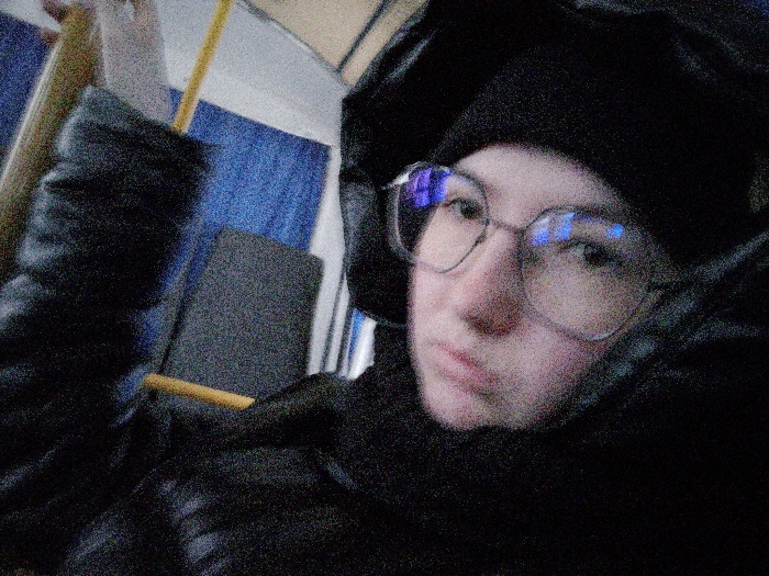 An individual is depicted in a close-up, low-light photograph taken inside what appears to be a public transport vehicle in Novyi Shagirt, Russia. The subject, who appears to be female, occupies the majority of the frame, looking directly towards the camera. She wears a dark, possibly black, beanie or knitted cap and large, light-rimmed eyeglasses with prominent blue reflections on the lenses. Her outerwear consists of a dark, puffy or quilted jacket/coat, with a dark scarf or high collar covering part of her lower face. Her expression is neutral.
The background elements are consistent with a passenger compartment. To the left and slightly behind the individual, a dark grey upholstered seat back is visible, featuring a horizontal wooden or light-colored bar, likely a handle or structural component. Further back, a textured blue surface, possibly upholstery or wall paneling, is discernible. A partial yellow vertical pole, common in public transport for standing passengers, is visible above the seat. The overall image exhibits significant digital noise, characteristic of a photograph taken in low light conditions.
